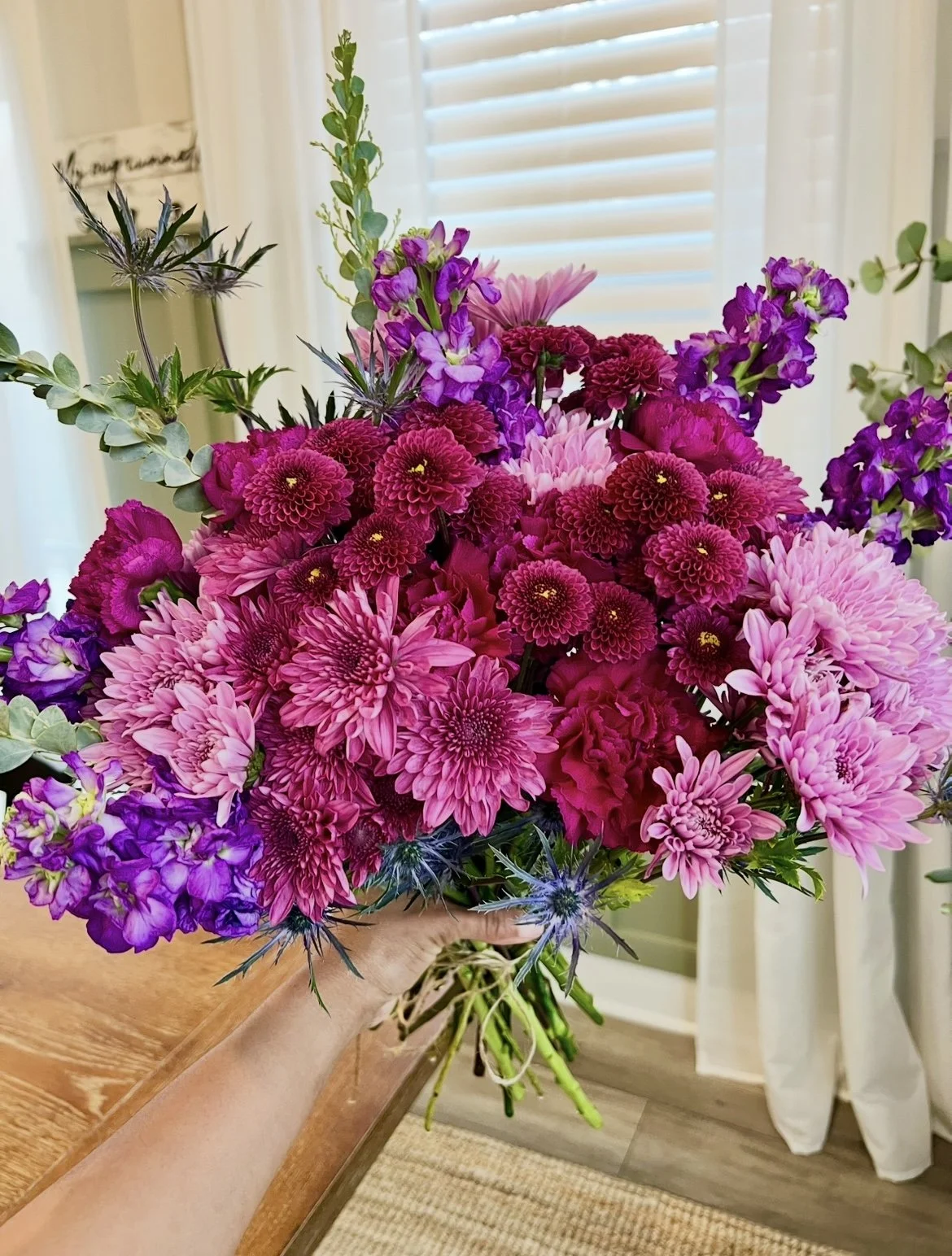 A person holding a vibrant bouquet of pink, purple, and magenta flowers inside a room with a window and white curtains.