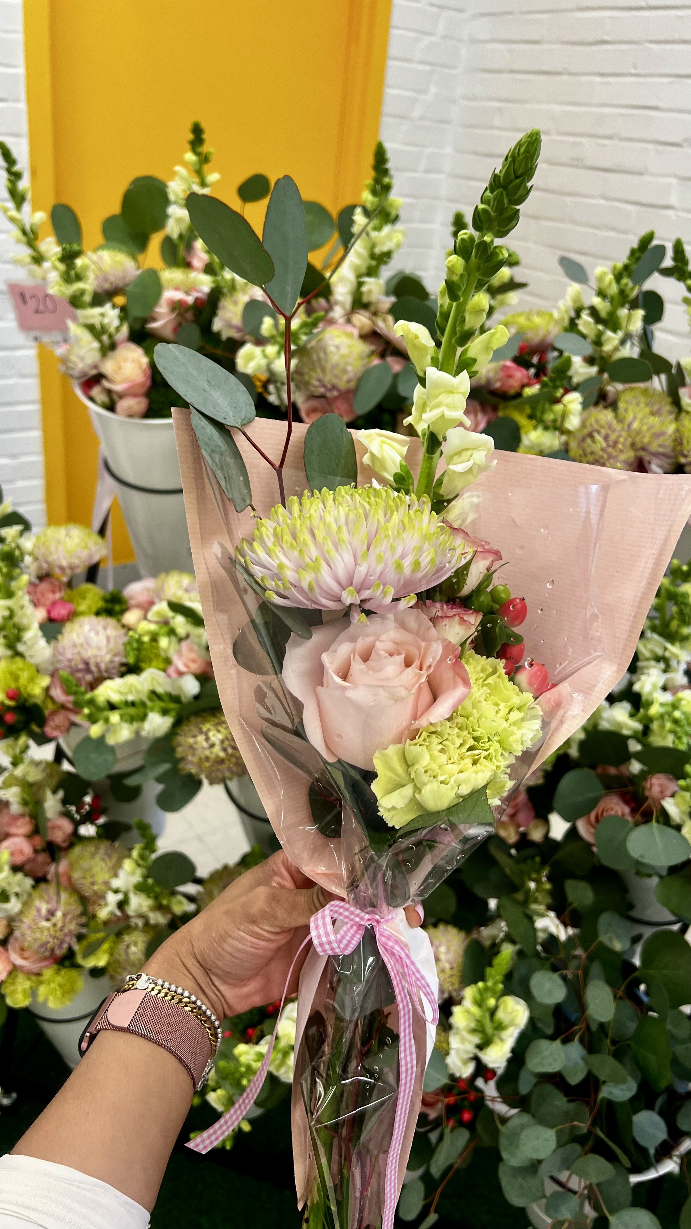 A hand holding a small bouquet of pink and white flowers wrapped in pink paper and tied with a pink gingham ribbon, in a flower shop with other floral arrangements in the background.