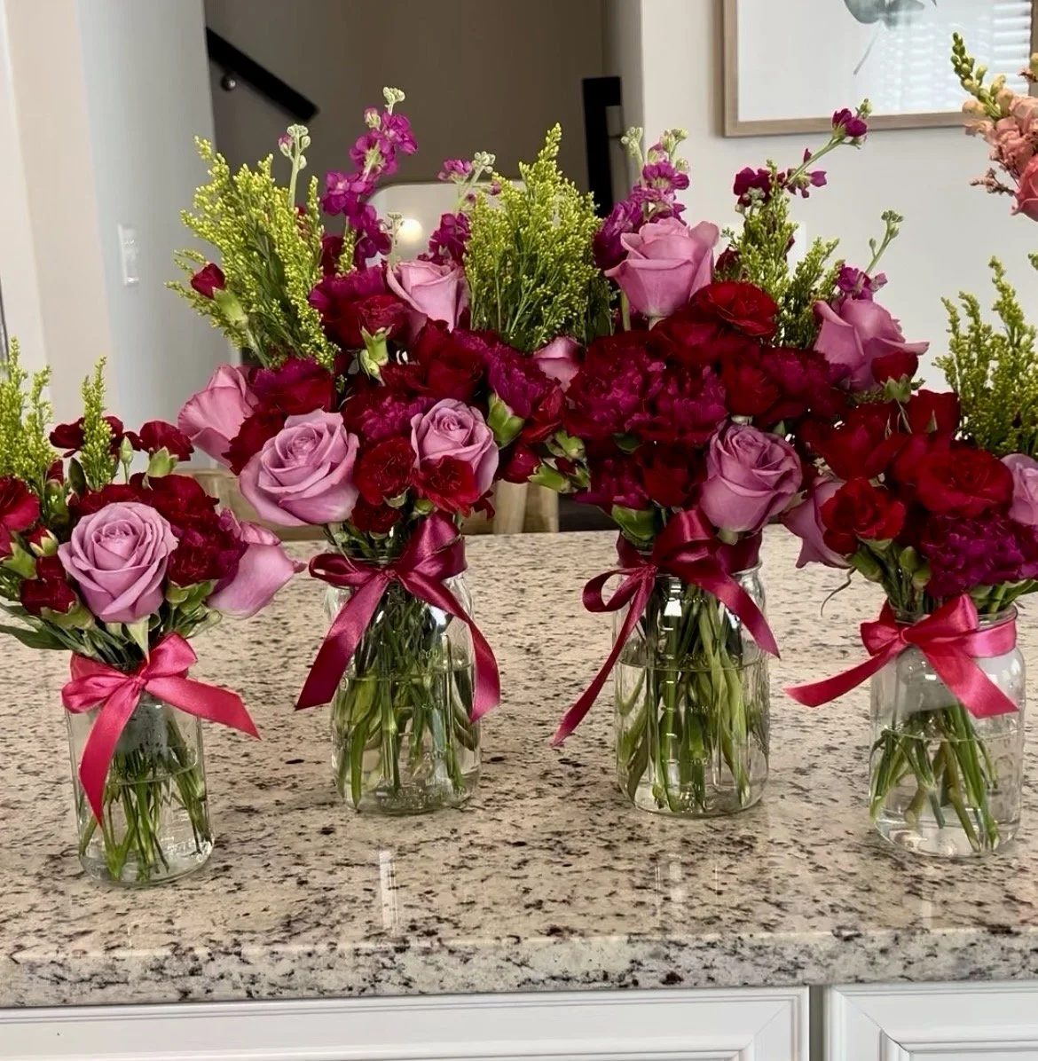 Four glass jars with bouquets of pink and red roses and greenery, each tied with a pink or red ribbon, placed on a granite countertop.
