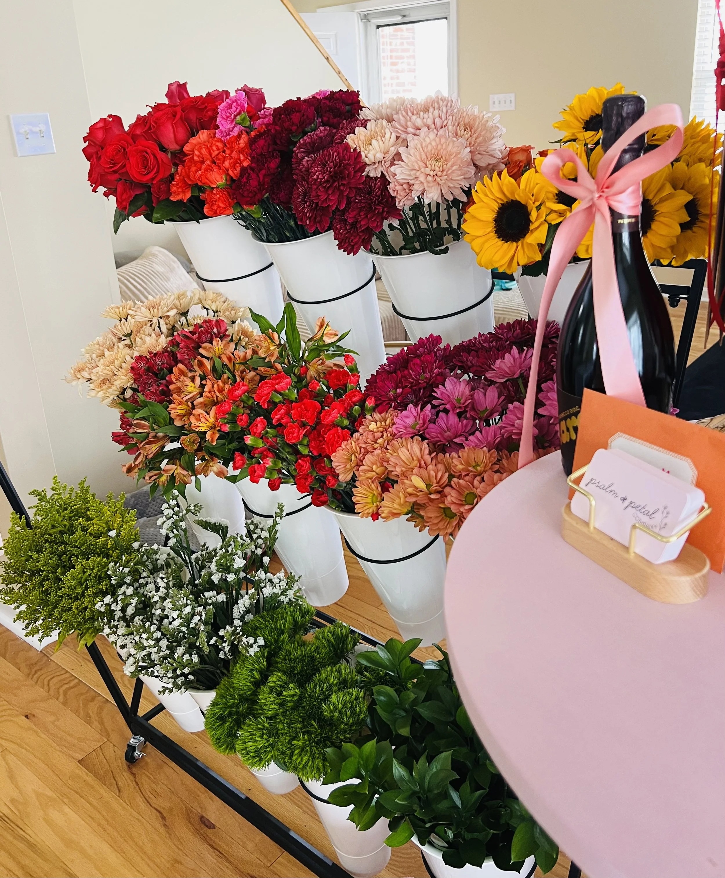 Various colorful flower arrangements and potted plants on display in a room, including roses, chrysanthemums, and greenery.