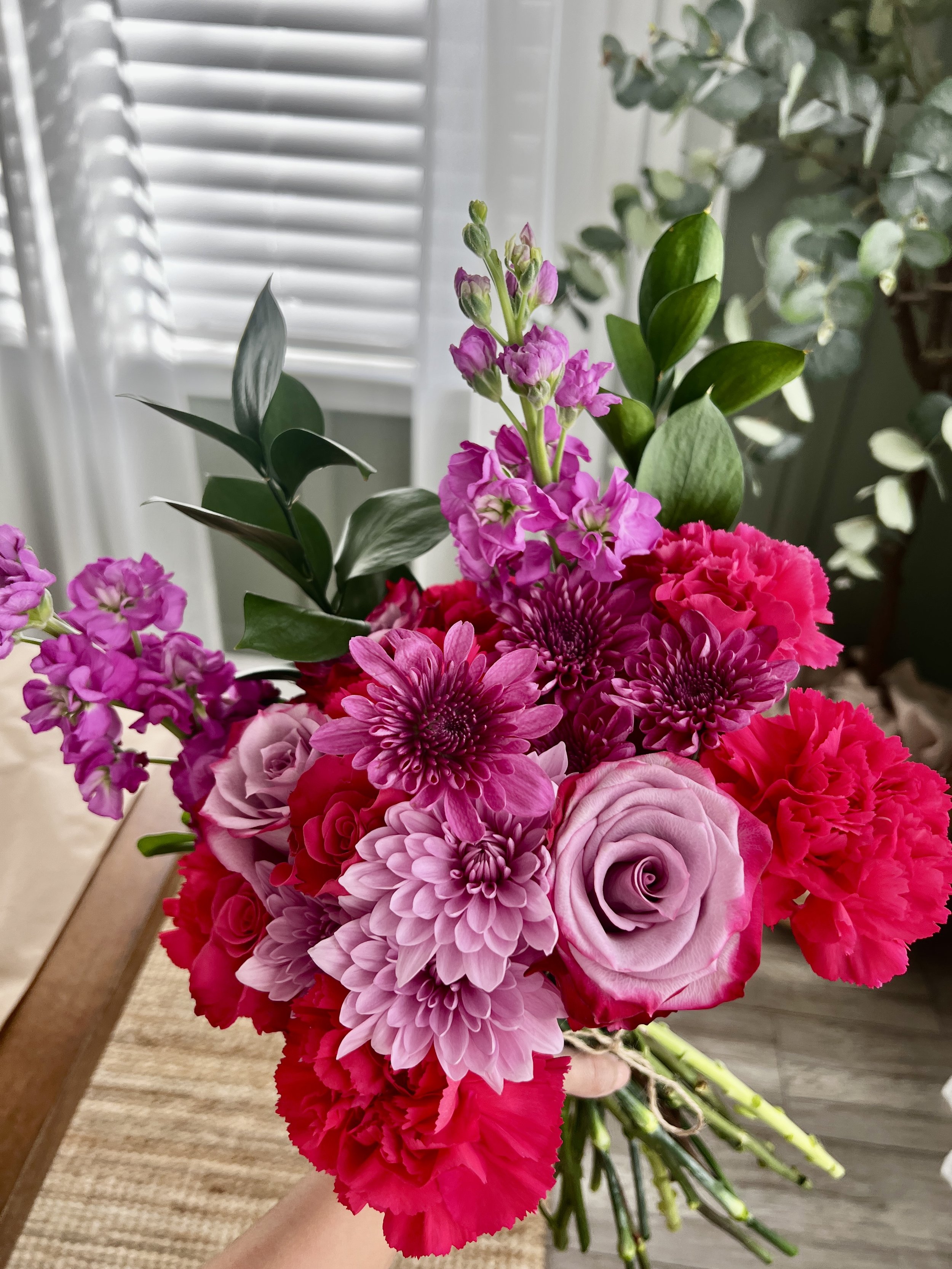 A bouquet of pink and purple flowers, including roses, dahlias, and stock, held by a hand indoors with a window and curtains in the background.