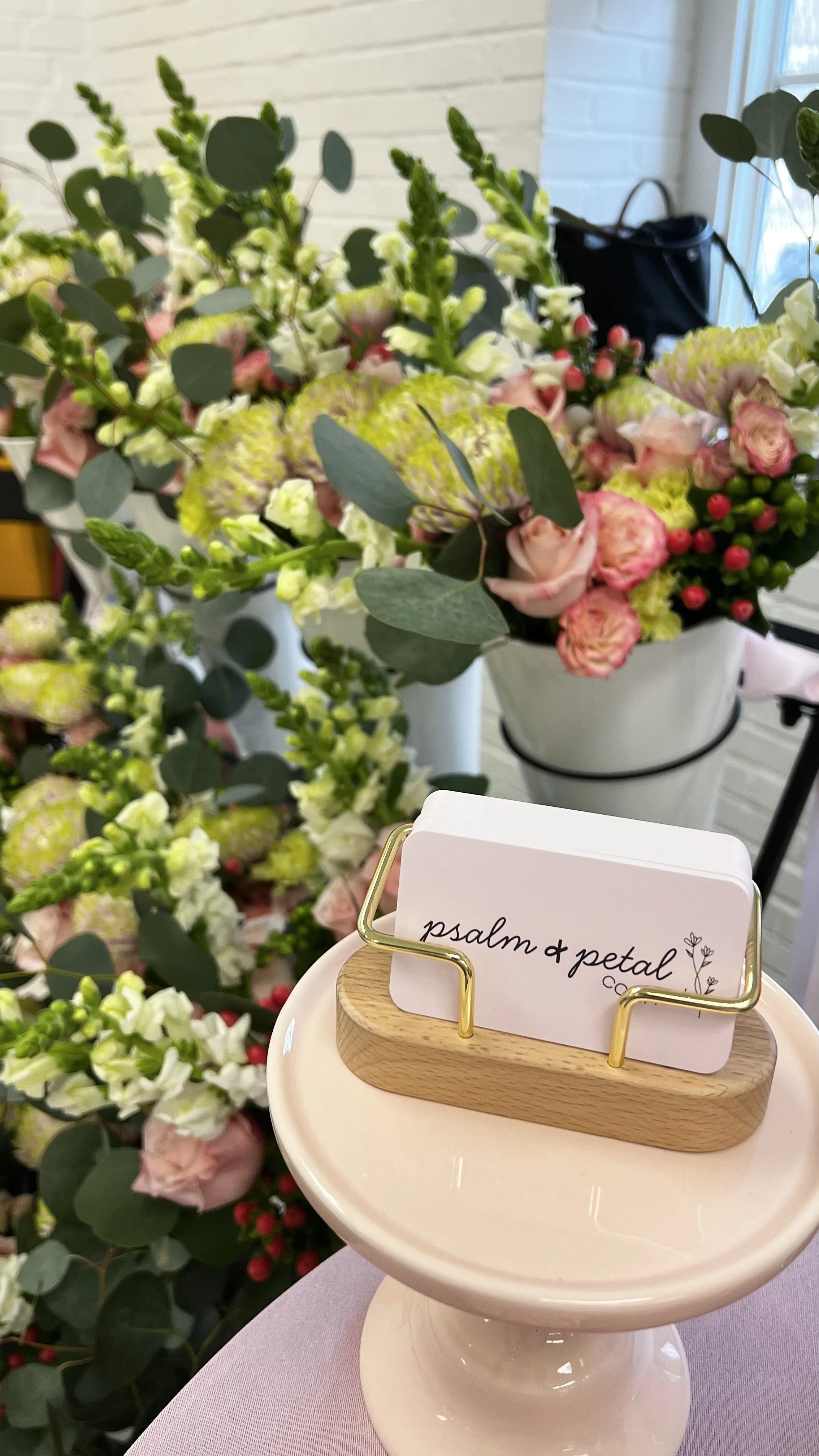 A flower arrangement with pink and white roses, green foliage, and other flowers in a white bucket. A small sign with 'psalm & petal' written on it is placed on a pink ceramic cake stand.