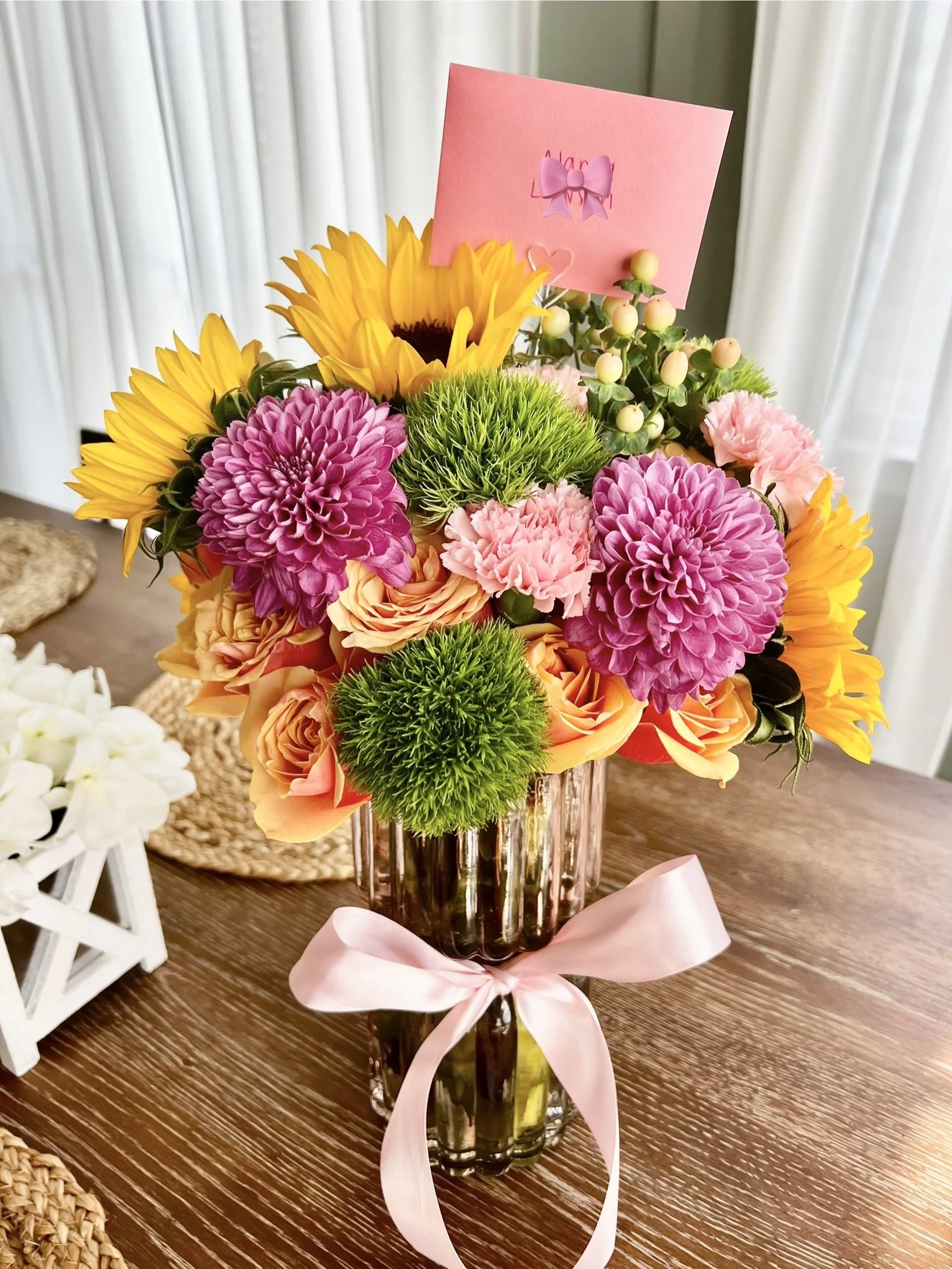 Colorful bouquet of sunflowers, pink dahlias, green chrysanthemums, and pink carnations in a glass vase with a pink ribbon, placed on a wooden table.