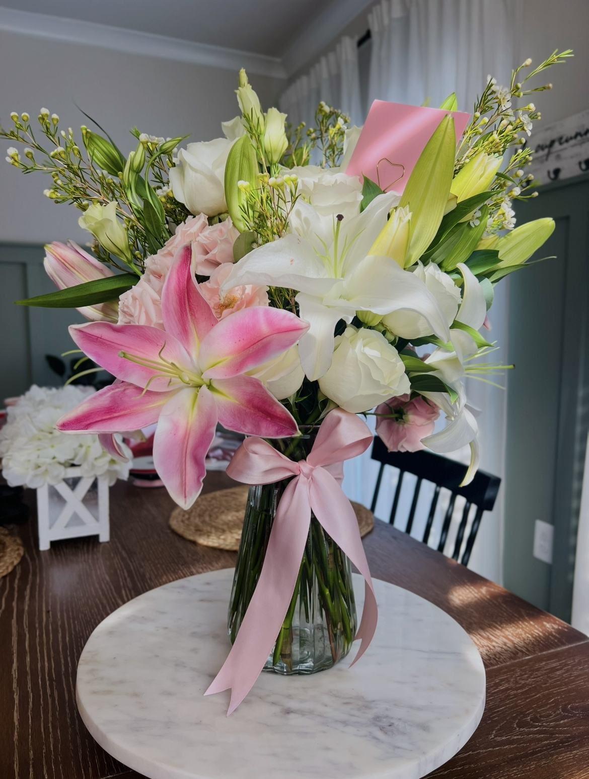 A glass vase with a pink ribbon bow holding a bouquet of white, pink, and yellow flowers on a white marble tray on a wooden table.