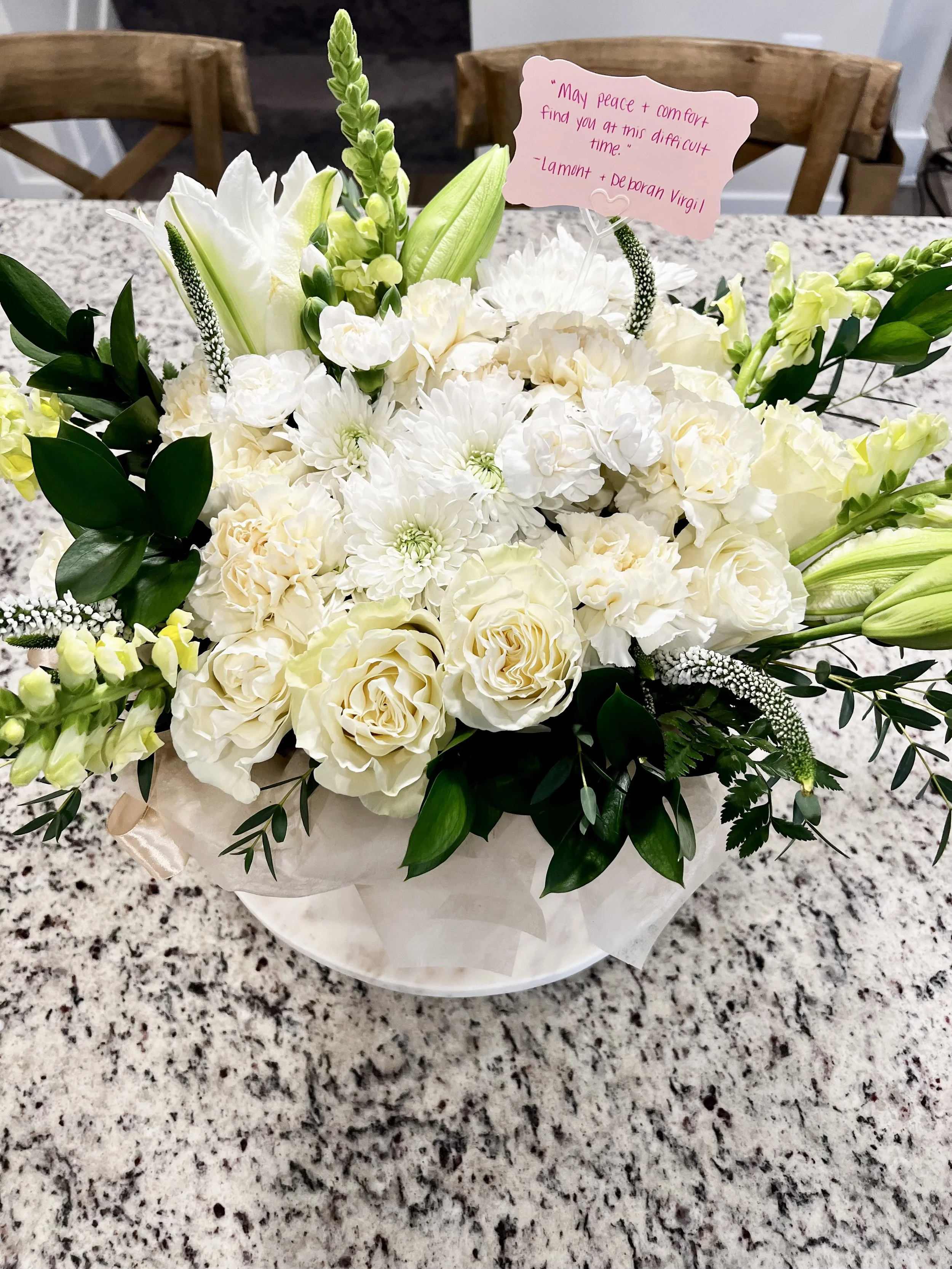 A floral arrangement of white roses, lilies, and other white flowers with green foliage, placed on a speckled granite countertop. A pink note card with a handwritten message is tucked into the flowers.
