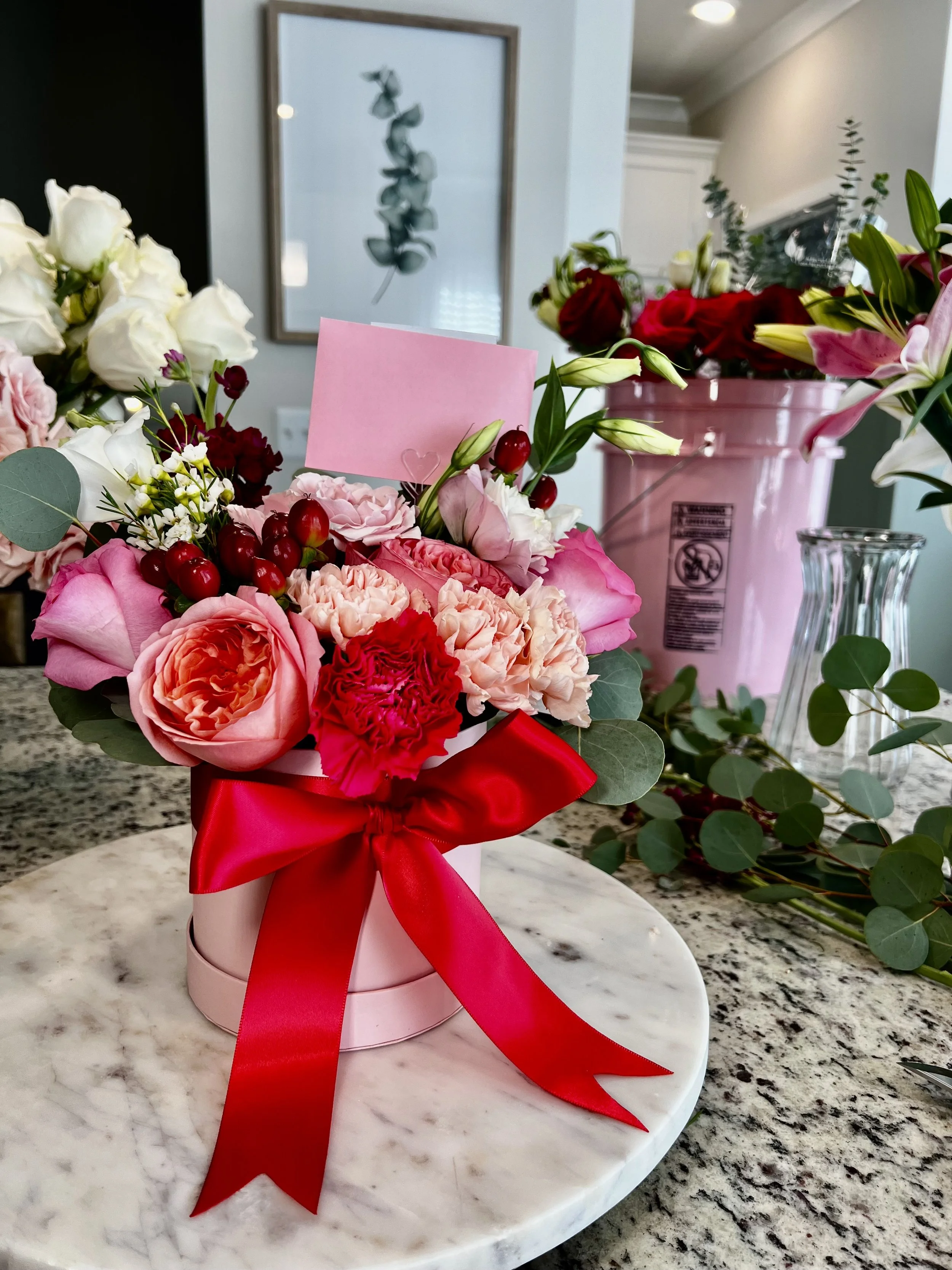 Pink flower bouquet with roses and carnations in a pink box with a red ribbon, placed on a marble surface.