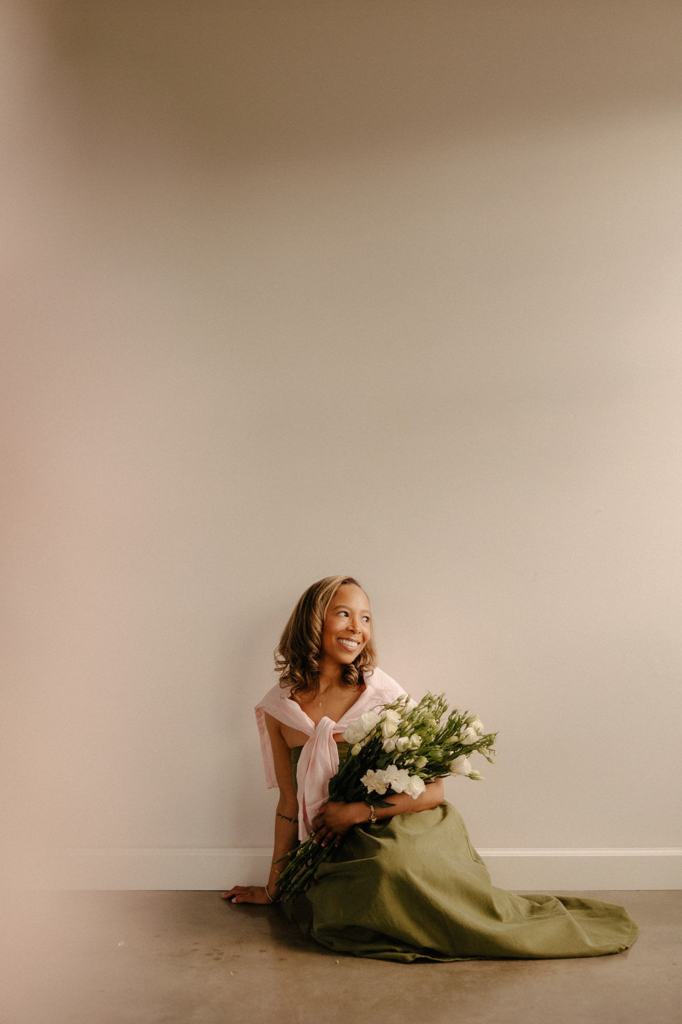 Girl in green dress holding white lisianthus bouquet while sitting on the floor gazing right.