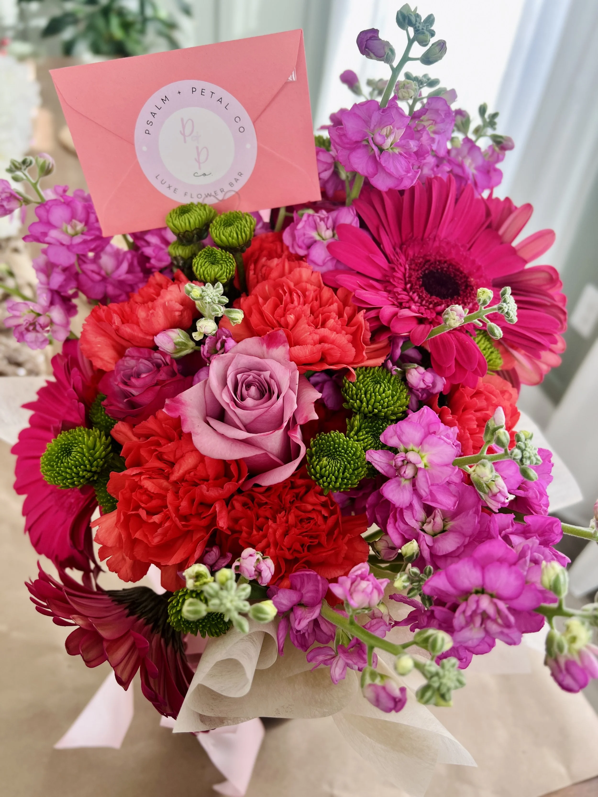 Bright pink, red, purple, and green mixed flower bouquet including roses, gerbera daisies, carnations, and other flowers with a pink card from Psalm + Petal Co attached.