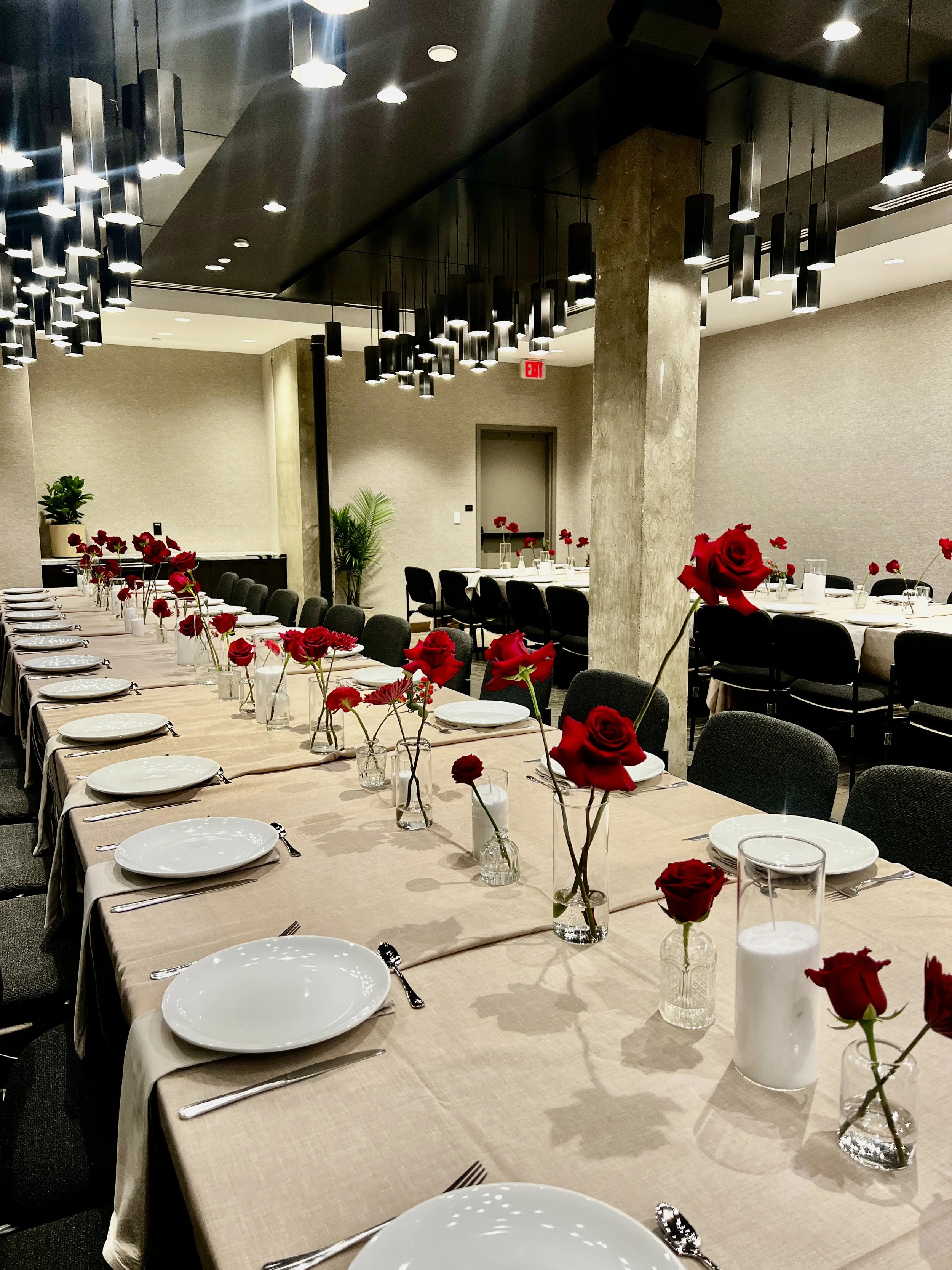 A banquet hall with a long table covered with a beige tablecloth, set with white plates, silverware, and glasses. Red roses in small glass vases are arranged along the table. Modern black lighting fixtures hang from the ceiling, and there are plants 
