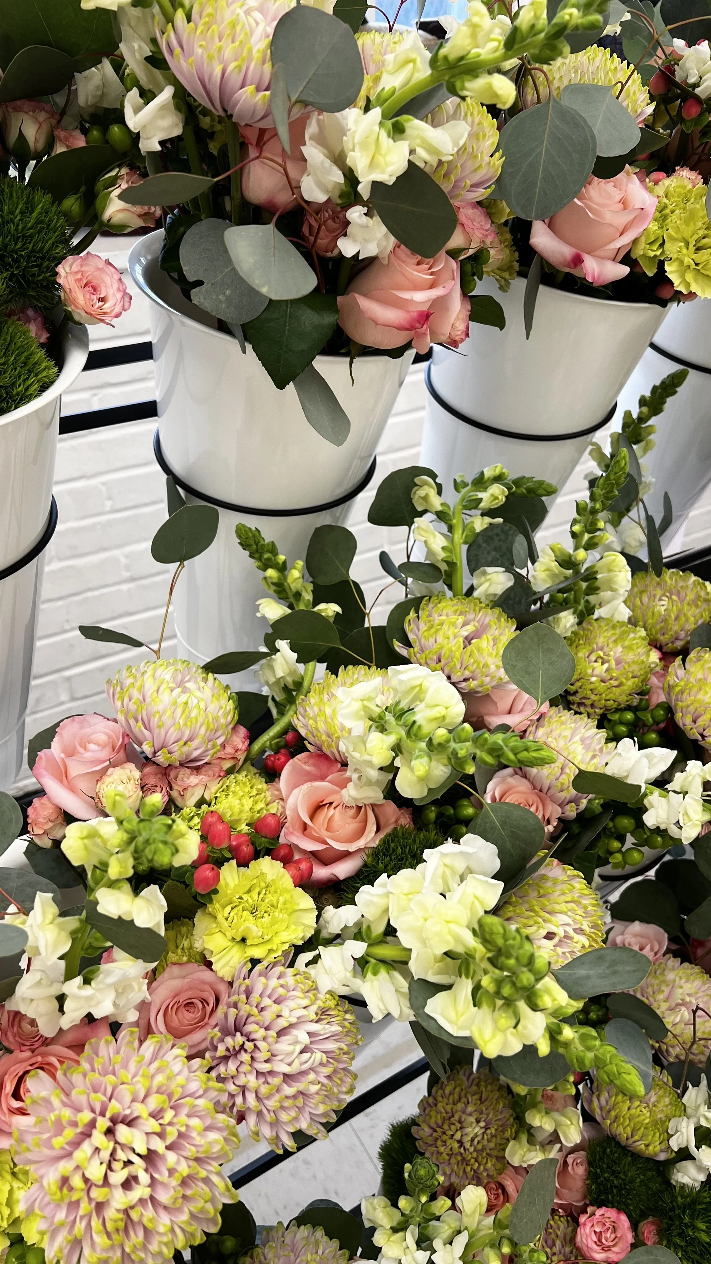 Displays an assortment of pink roses, light green chrysanthemums, white snapdragons, greenery, and red berries arranged in white buckets against a white brick background.