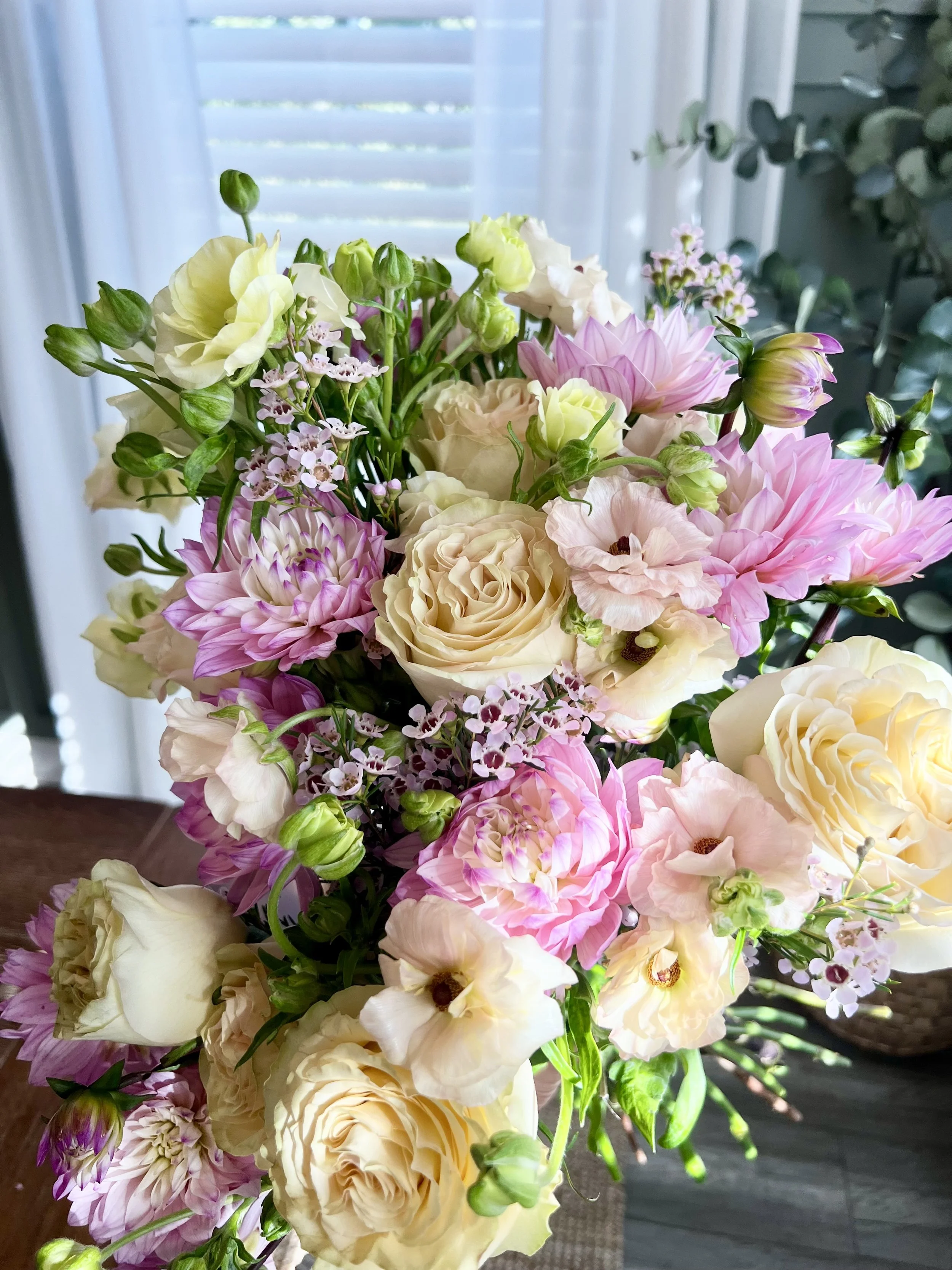 A bouquet of mixed flowers including roses, dahlias, and lisianthus in soft pastel colors, set against a background of a window with blinds.