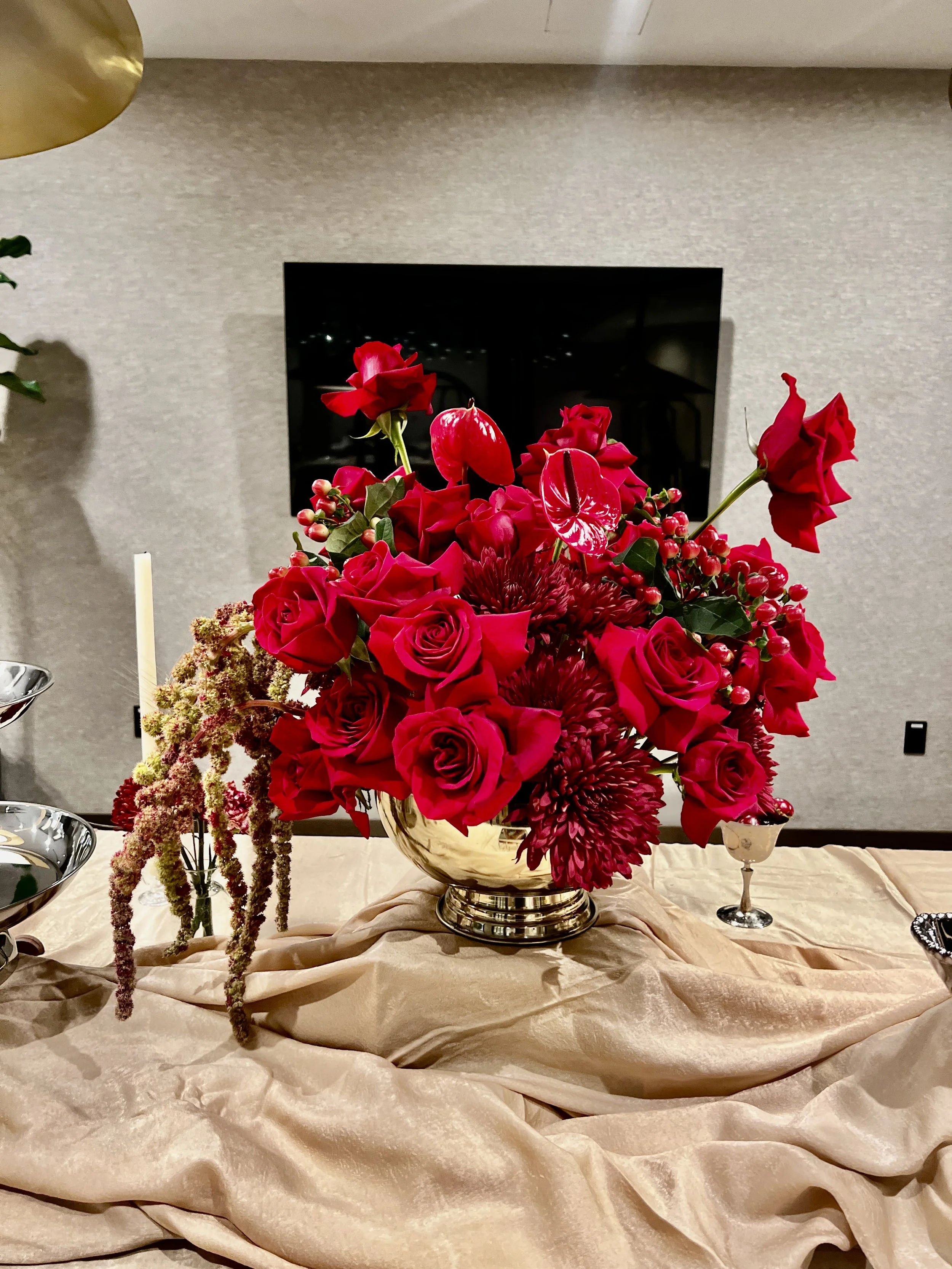 A large floral arrangement with red roses, red anthuriums, and burgundy dahlias in a shiny gold vase on a table with a beige tablecloth.