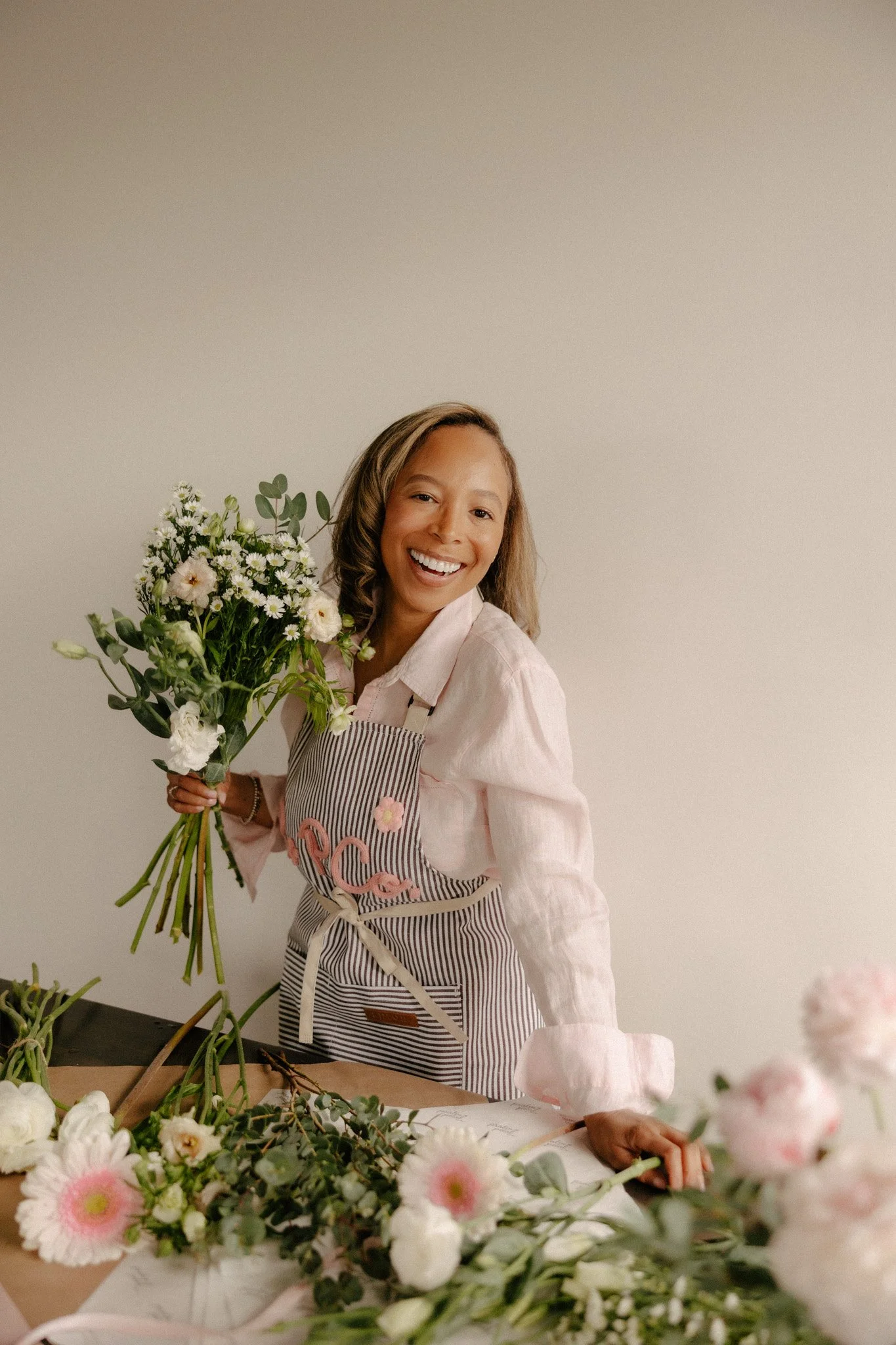 Girl leaning over table of flowers smiling in an apron while holding a white bouquet.