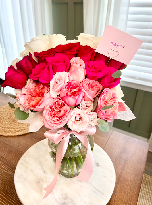 A bouquet of pink, red, and white roses in a glass vase with a pink ribbon, accompanied by a note that says 'madre' with a heart, on a wooden table near a window with white blinds.