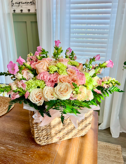 A basket of pink, white, and green flowers on a wooden table, with white curtains and window blinds in the background.