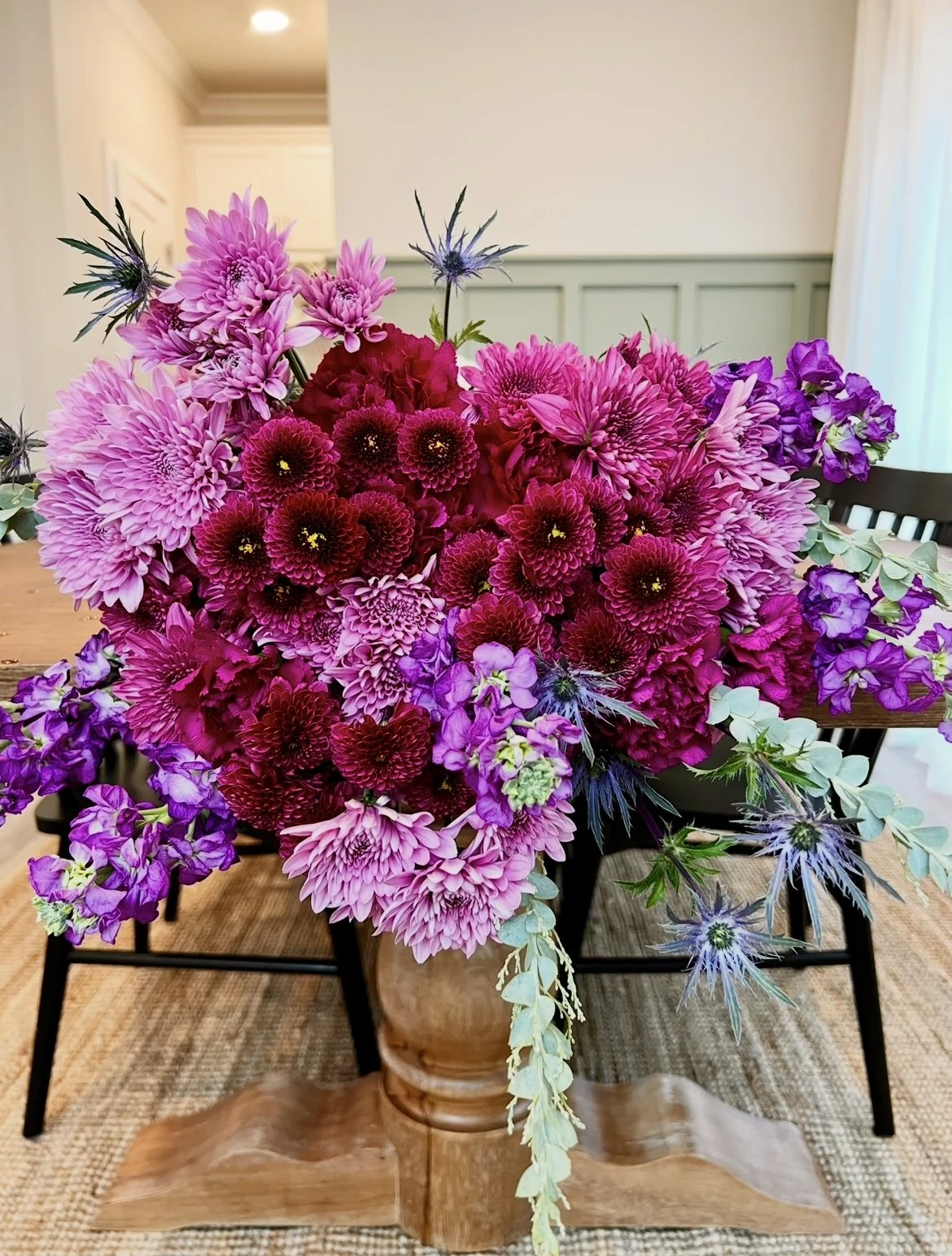 A large colorful bouquet of pink, purple, and red flowers in a tan vase on a wooden table in a room with beige walls and a chair.