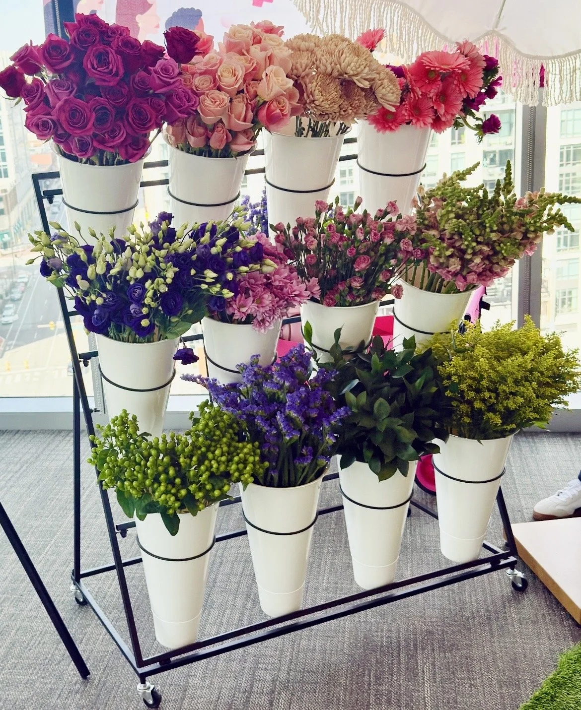 Multiple white cones filled with colorful flowers arranged on a tiered display stand near a large window with cityscape view.