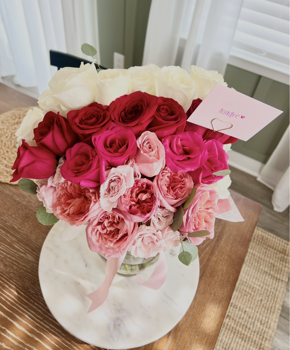A bouquet of pink, red, and white roses with green leaves in a glass vase on a wooden table, with a pink ribbon wrapped around the vase and a card that reads 'madre' in pink ink attached with a gold paperclip.