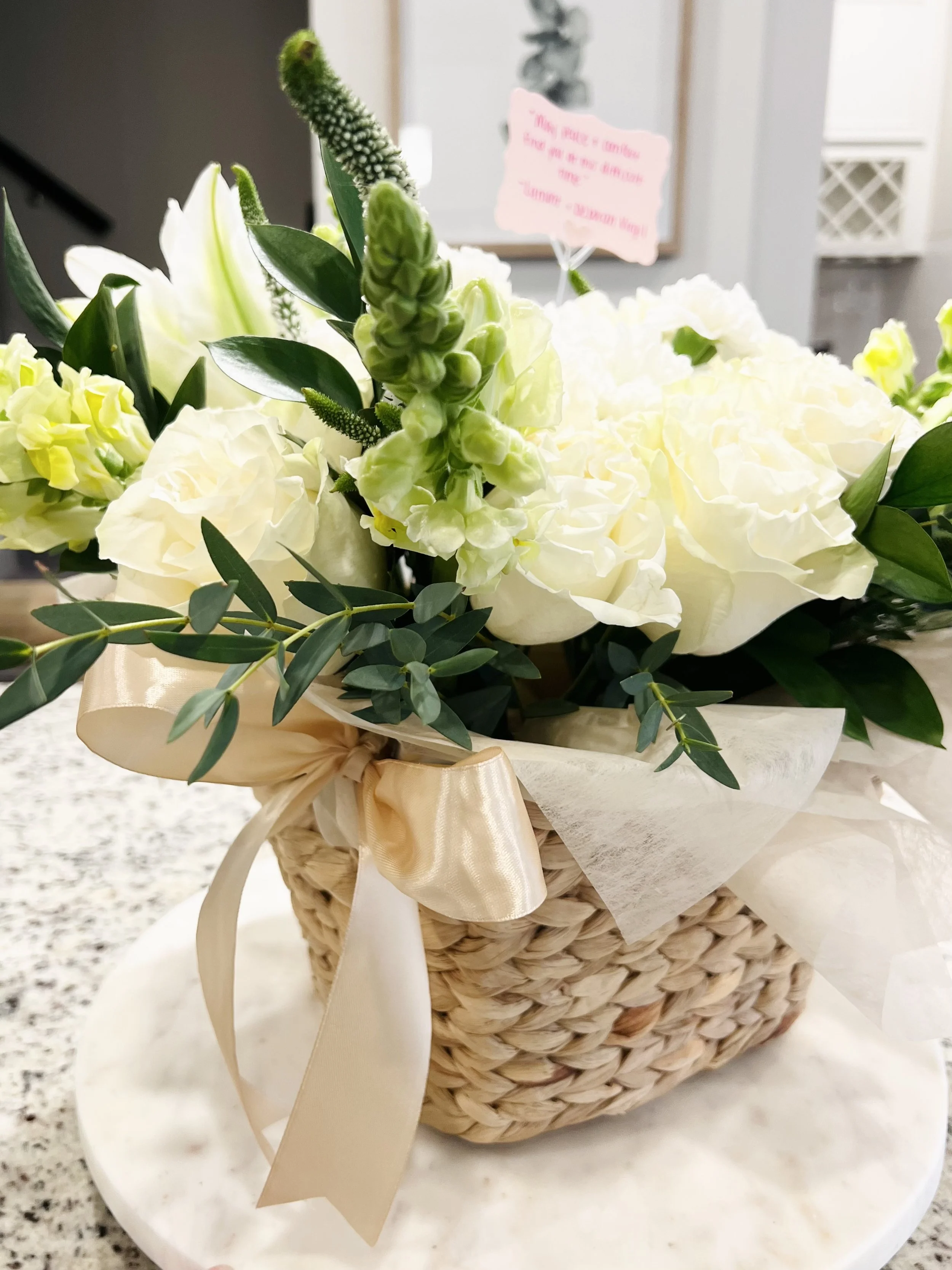 A white flower arrangement with greenery and a beige bow in a woven basket, placed on a marble surface.