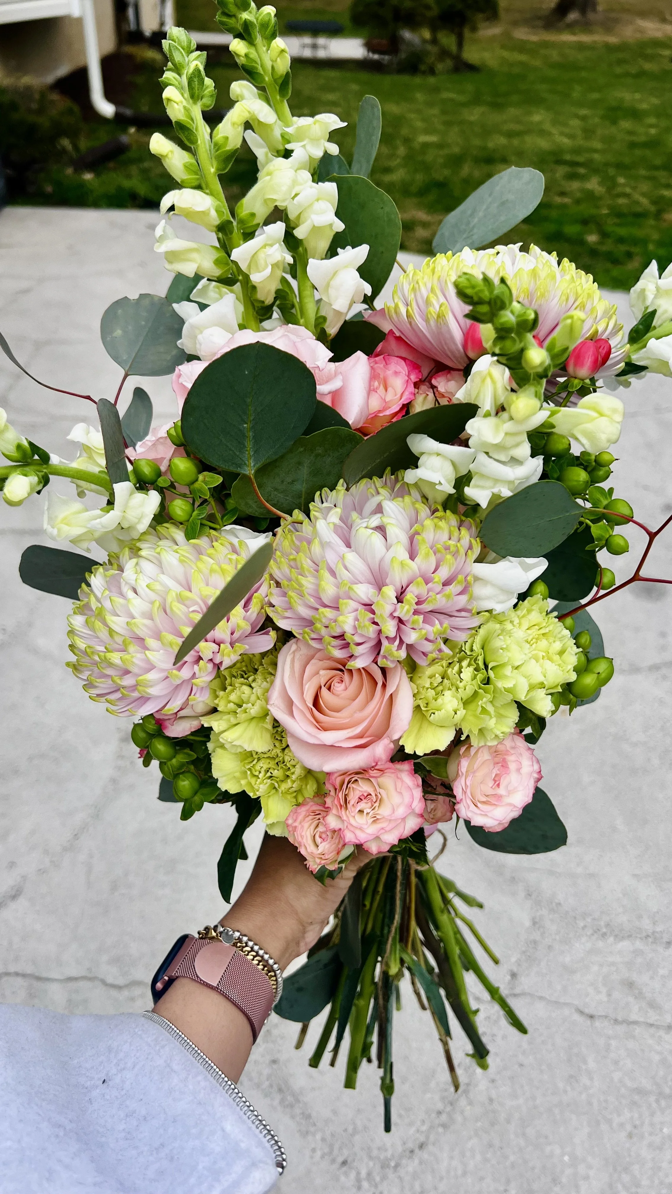 A hand holding a colorful bouquet of pink, white, and green flowers with greenery in an outdoor setting.