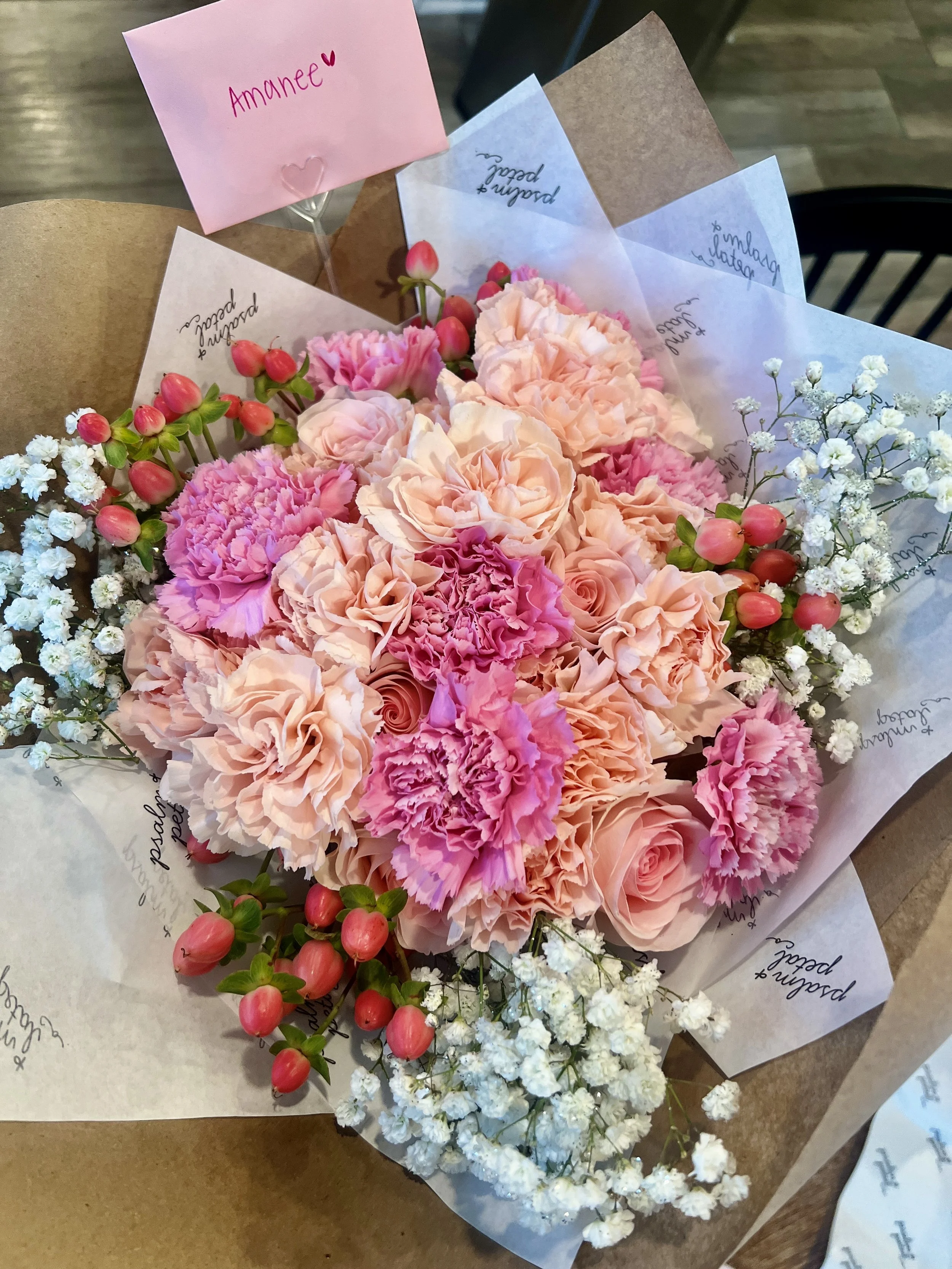 Pink and white flower bouquet with roses, carnations, baby's breath, and hypericum berries, wrapped in brown paper and white parchment paper, with a pink note that says 'Amanée' in the background.