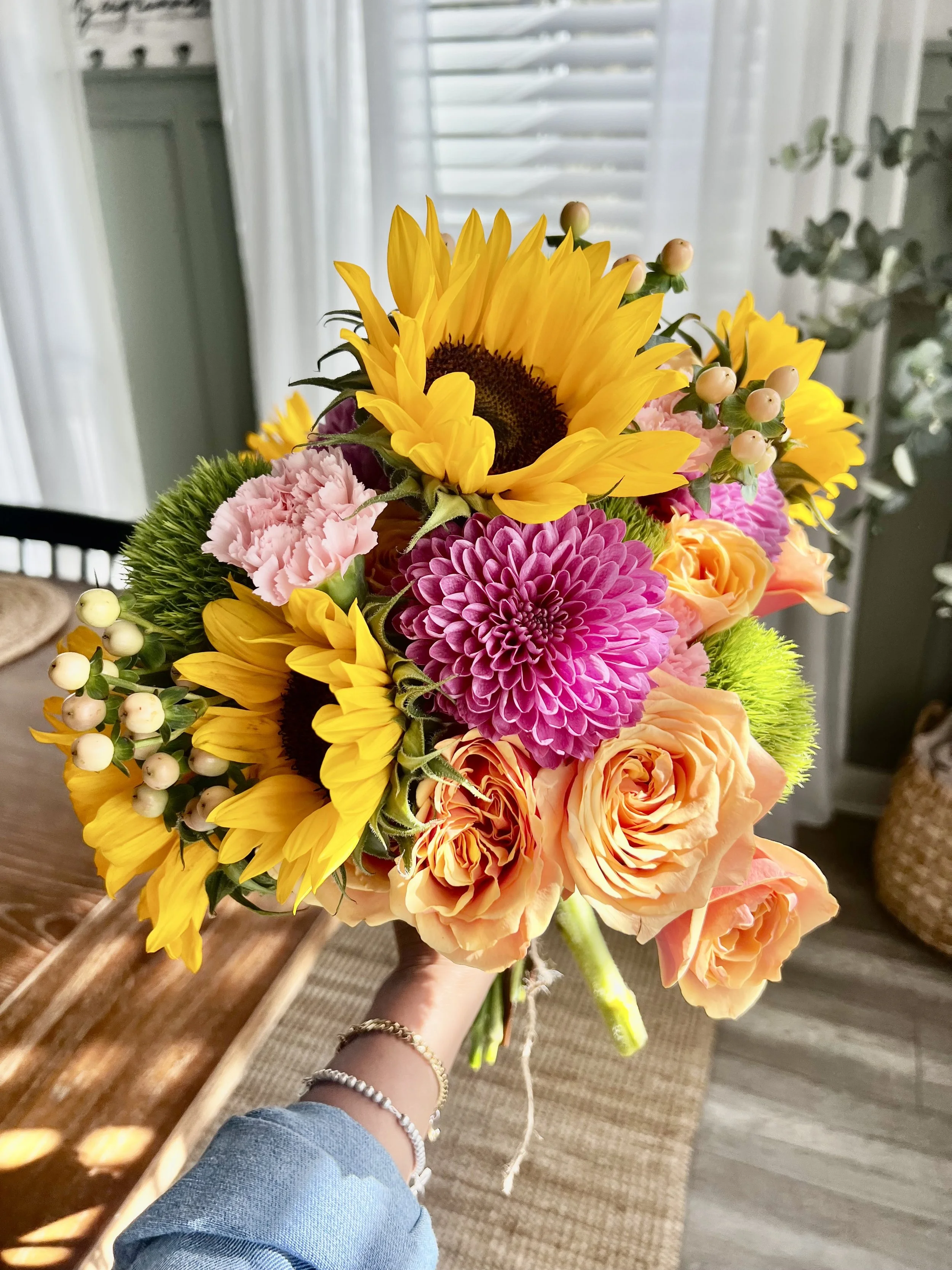 Flower bouquet with sunflowers, pink dahlias, peach roses, pink carnations, and white berries held by a person wearing silver bracelets.