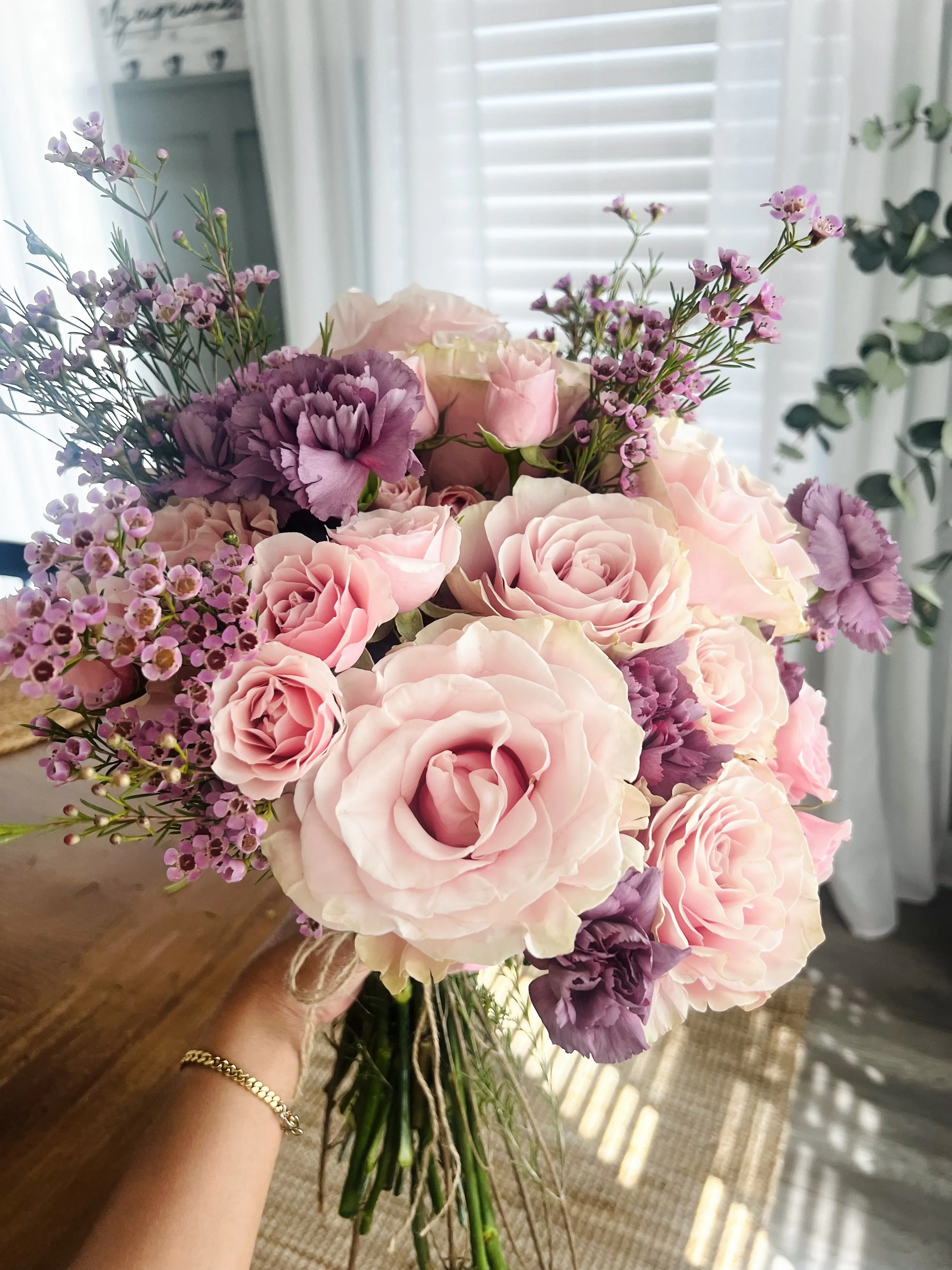 A hand holding a bouquet of pink and purple flowers, including roses and baby's breath, in a bright room with white blinds in the background.