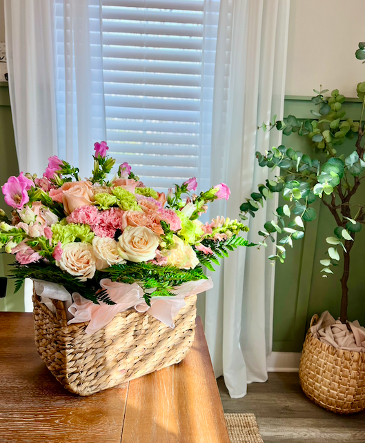 A basket of pink, cream, and green flowers on a wooden table near a window with white blinds, and a potted plant to the right with greenery and a wicker basket