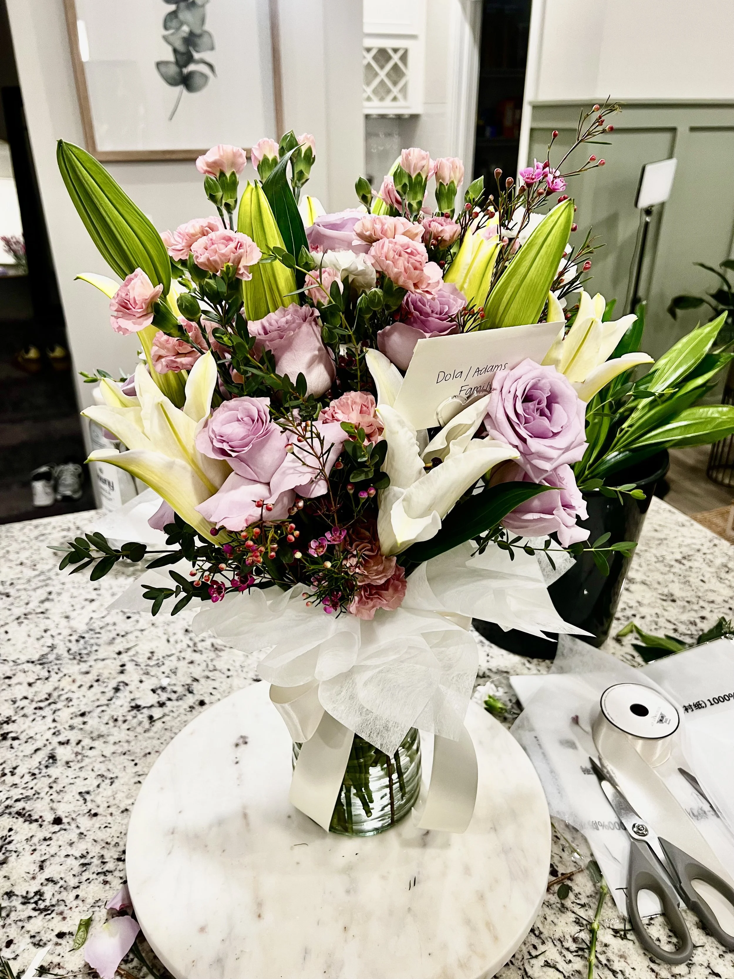 A tall bouquet of pink and white flowers, including lilies, roses, and small filler flowers, arranged in a vase on a marble table in a living room.