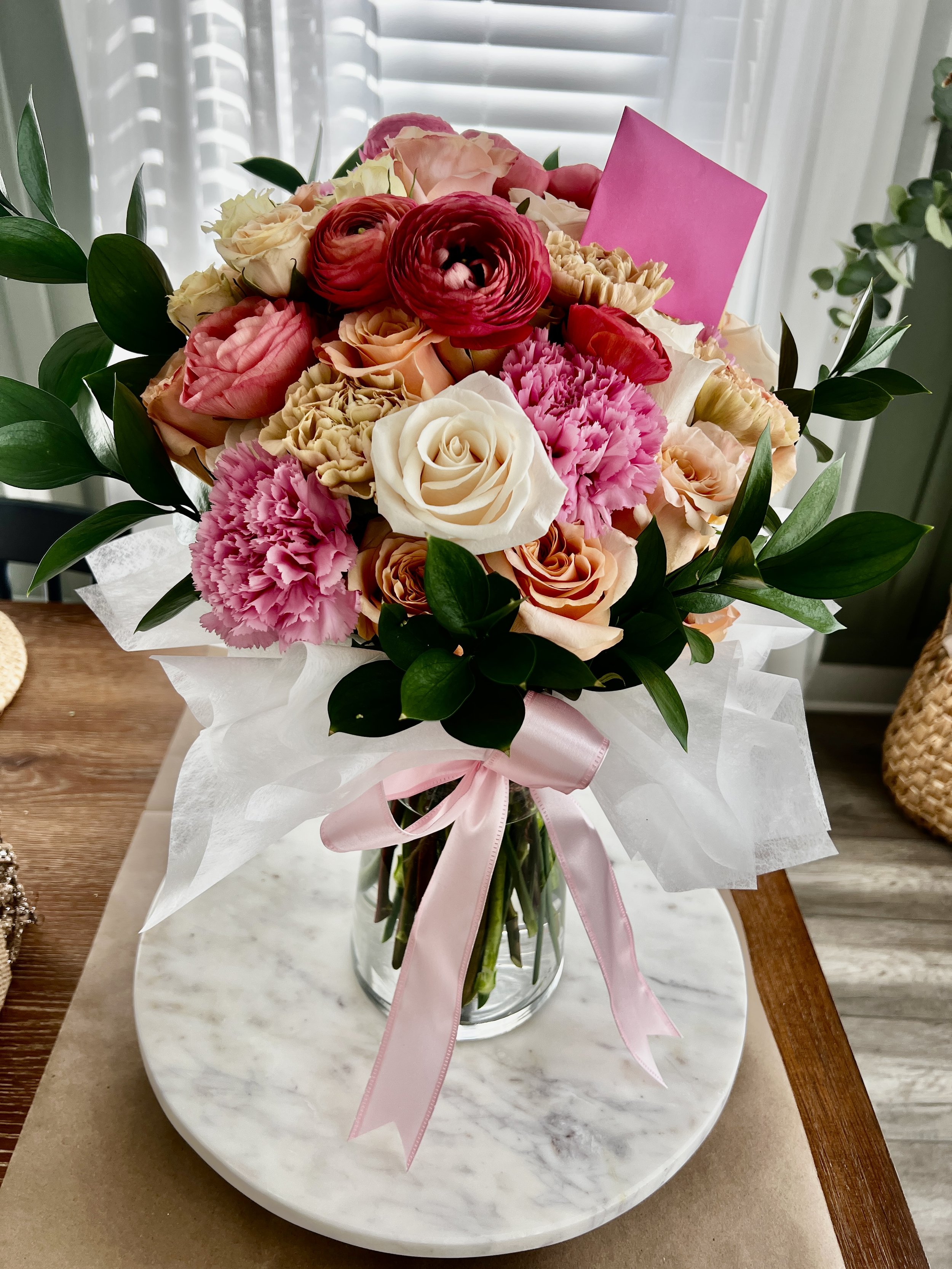 A bouquet of pink, white, and peach flowers arranged in a clear glass vase with a pink ribbon, placed on a white marble tray on a wooden table.