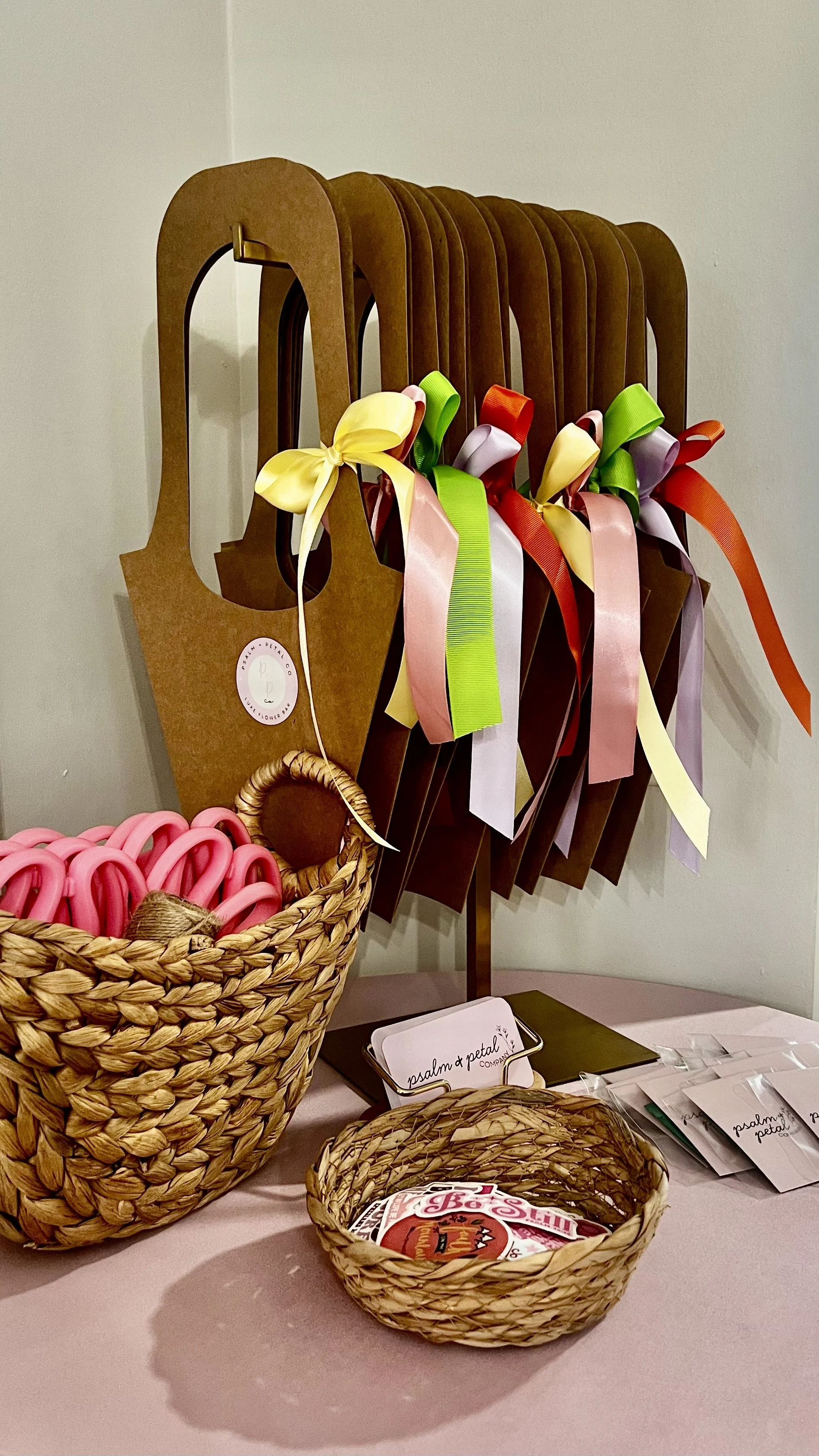 Decorative paper chain bows hanging on a display rack with a pink table, woven baskets with pink ribbon, business cards, and informational flyers.