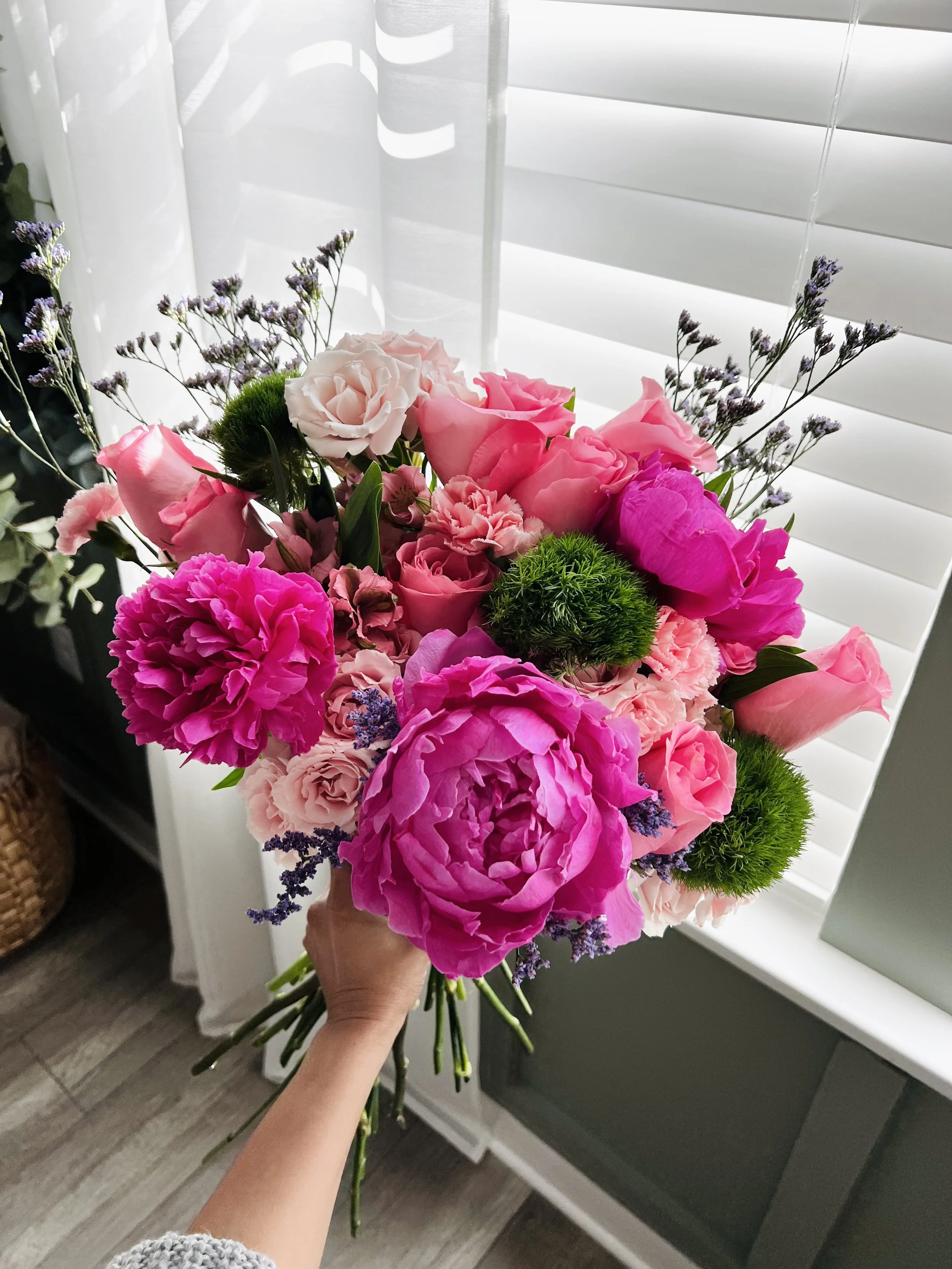 A hand holding a bouquet of pink and purple flowers in front of white window blinds.