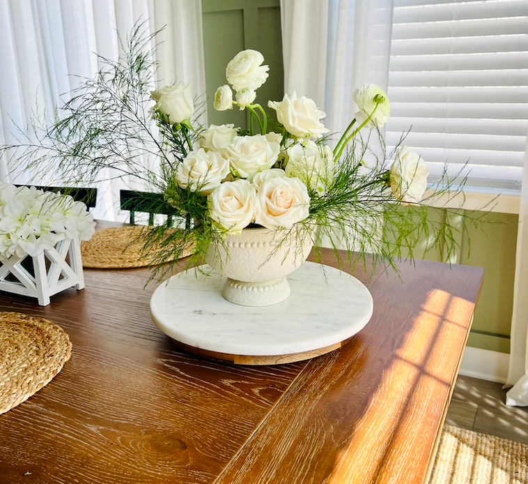 A floral arrangement of white roses and green foliage in a decorative white ceramic vase on a round marble tray on a wooden table.