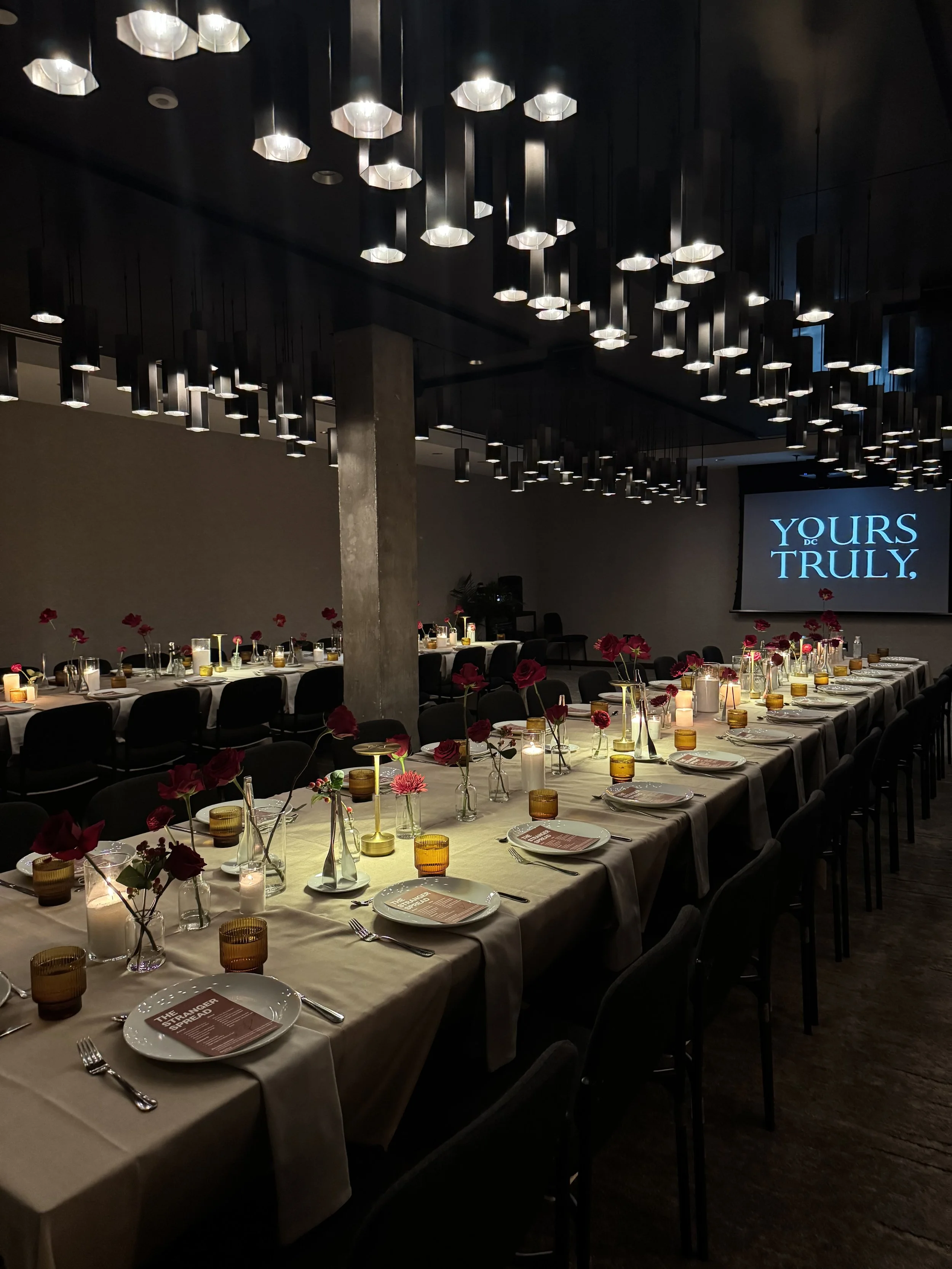 A long dinner table set for a formal event with candles, flowers, plates, glasses, and cutlery in a dimly lit room with modern pendant lighting and a projection screen displaying the words "YOURS TRULY."