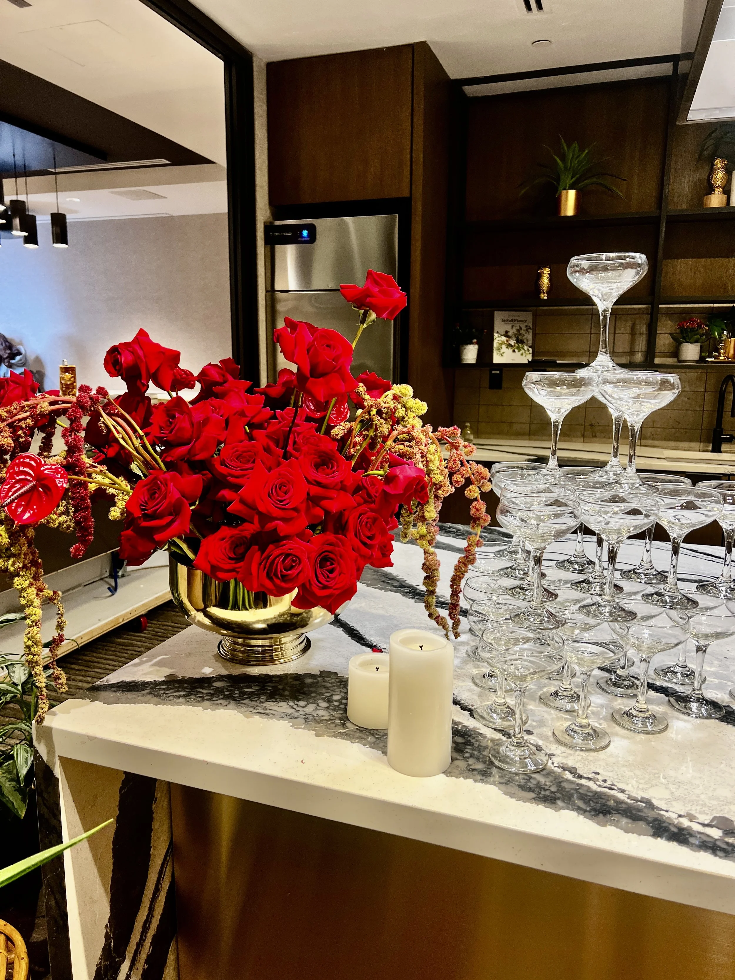 A bouquet of red roses and other flowers in a metallic vase on a marble countertop, with stacked empty cocktail glasses and two white candles nearby in a modern kitchen or bar area.