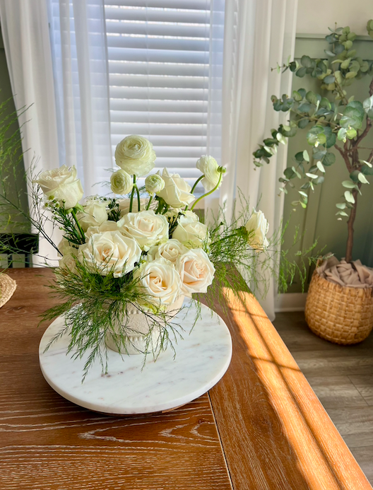 A white floral arrangement with white roses and ranunculus in a marble vase on a wooden table near a window with blinds, with a green potted plant in the background.