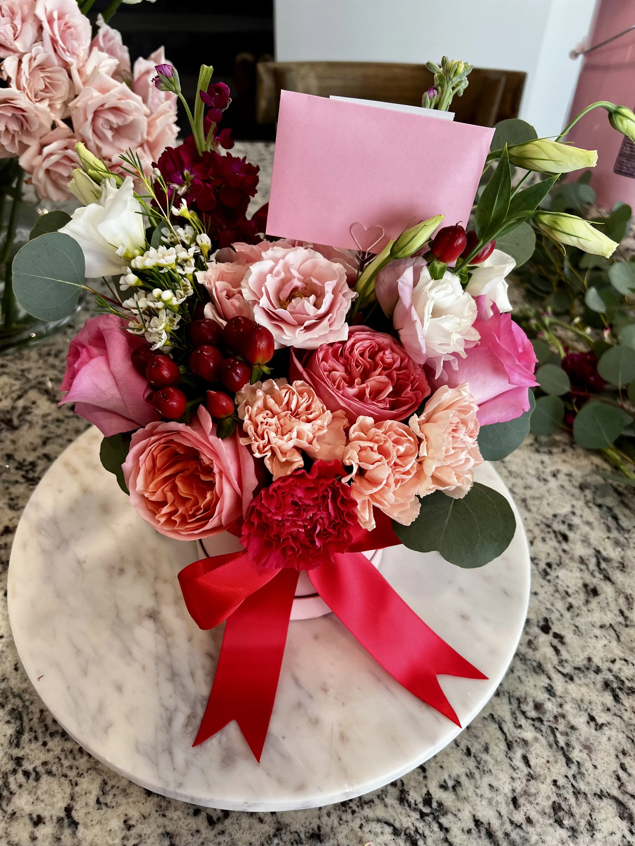 A bouquet of pink, red, and white flowers with green leaves and berries, decorated with a red ribbon and a pink card, placed on a white marble surface.