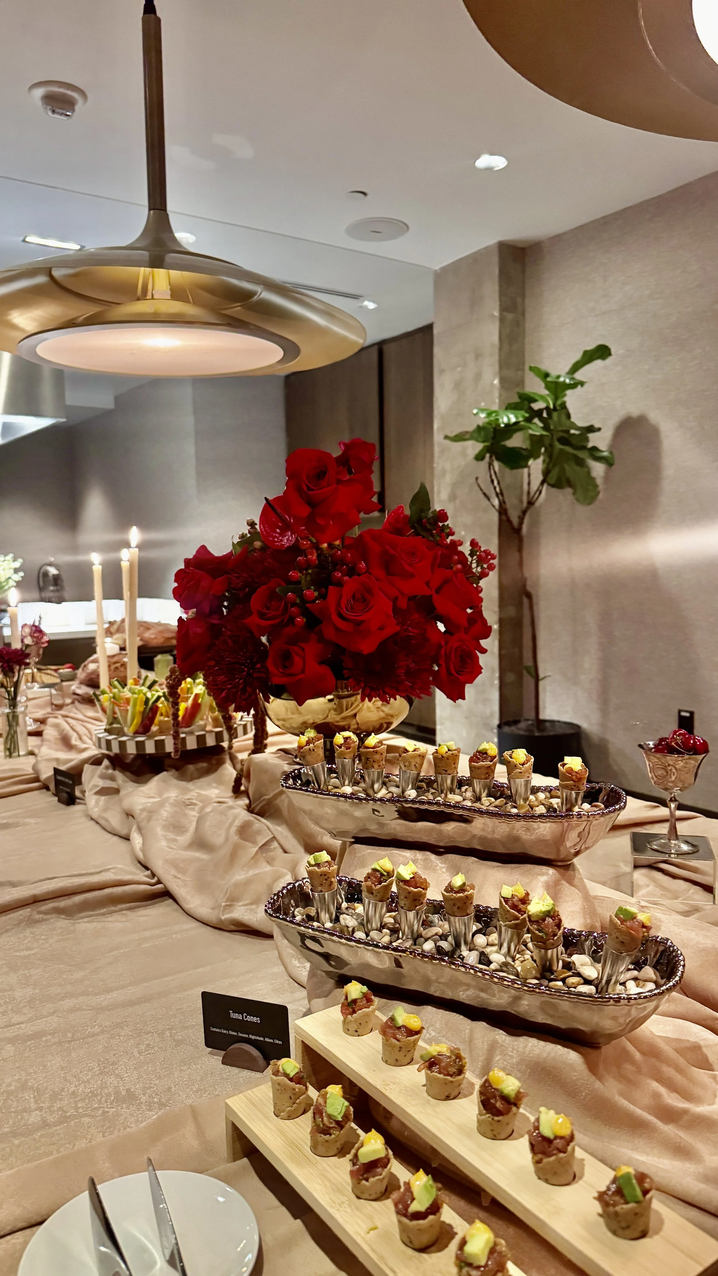 A buffet table decorated with a beige tablecloth, featuring a large bouquet of red roses, small plates of appetizers, and a dish of cherries in a decorative glass container. The setting appears to be an elegant dining area with modern lighting and a 
