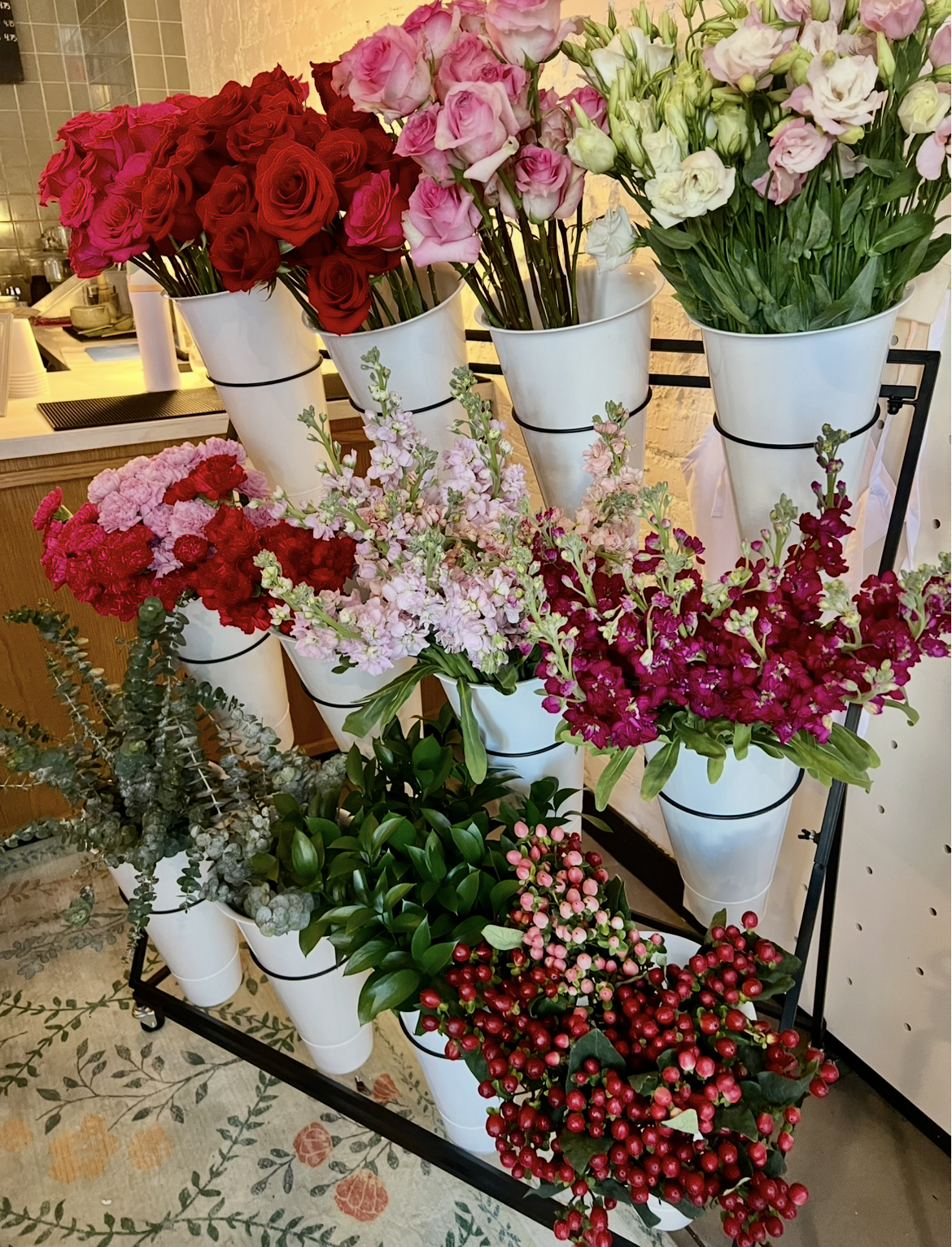 Various colorful flowers including pink, red, and white roses, pink snapdragons, and berries arranged in white vases on a display stand.