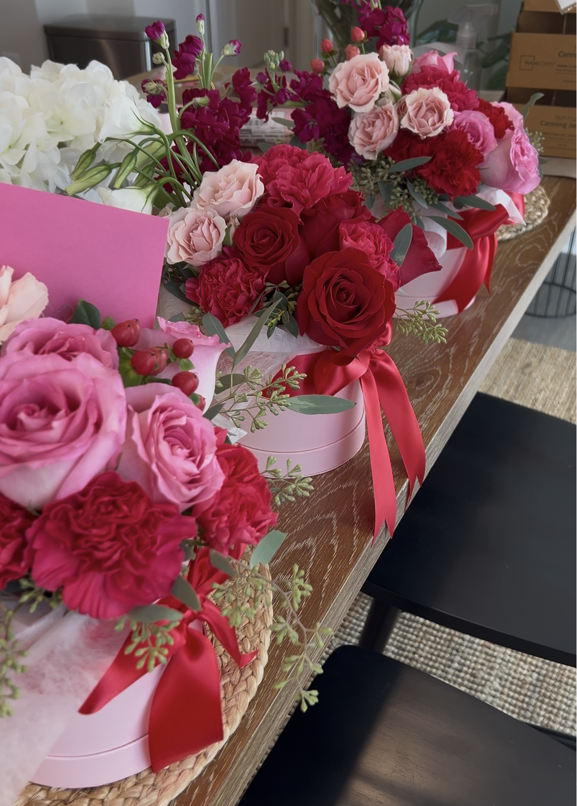 Multiple bouquets of pink and red roses, carnations, and other flowers with green foliage, arranged in pink and white boxes with red ribbons, displayed on a wooden table.