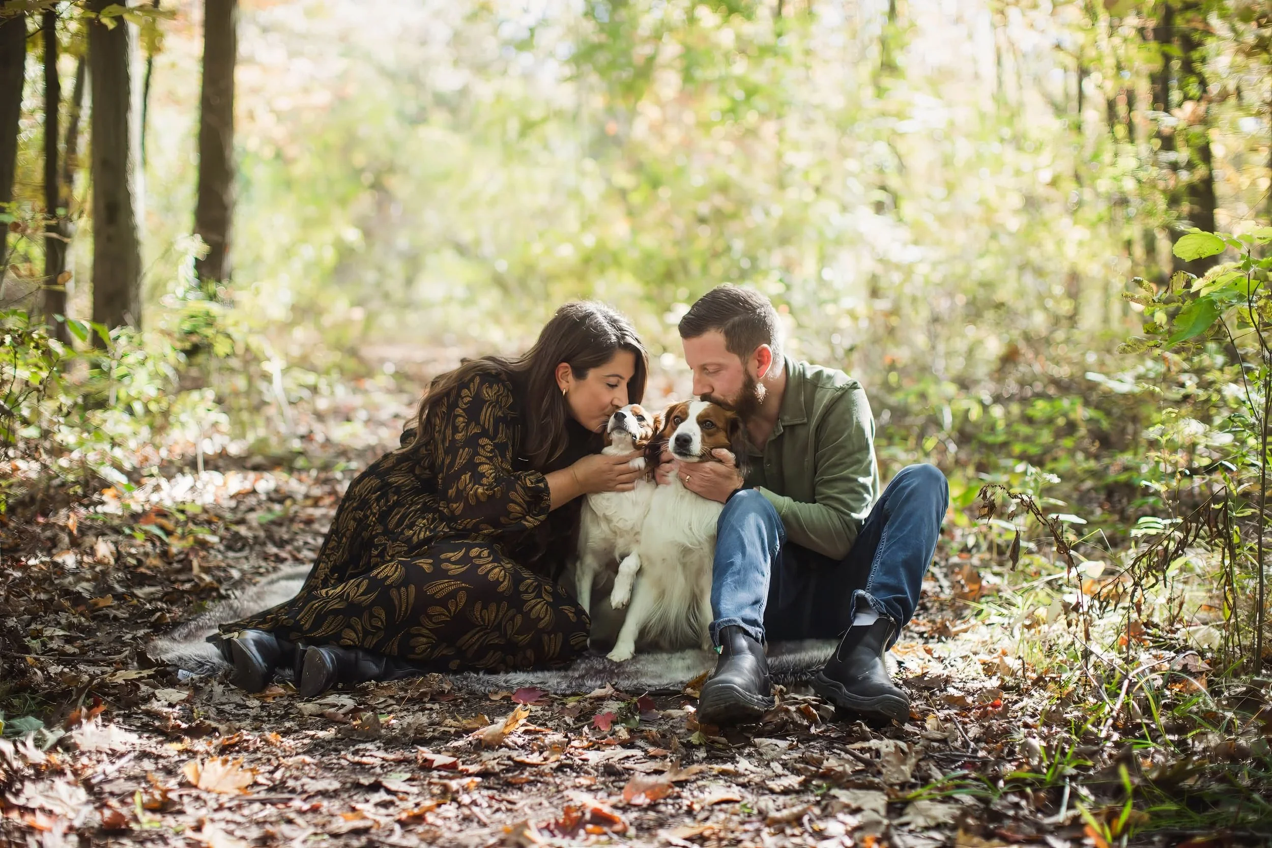 A couple sitting on a forest floor with their two Nederlandse Kooikerhondjes, surrounded by trees and autumn leaves.