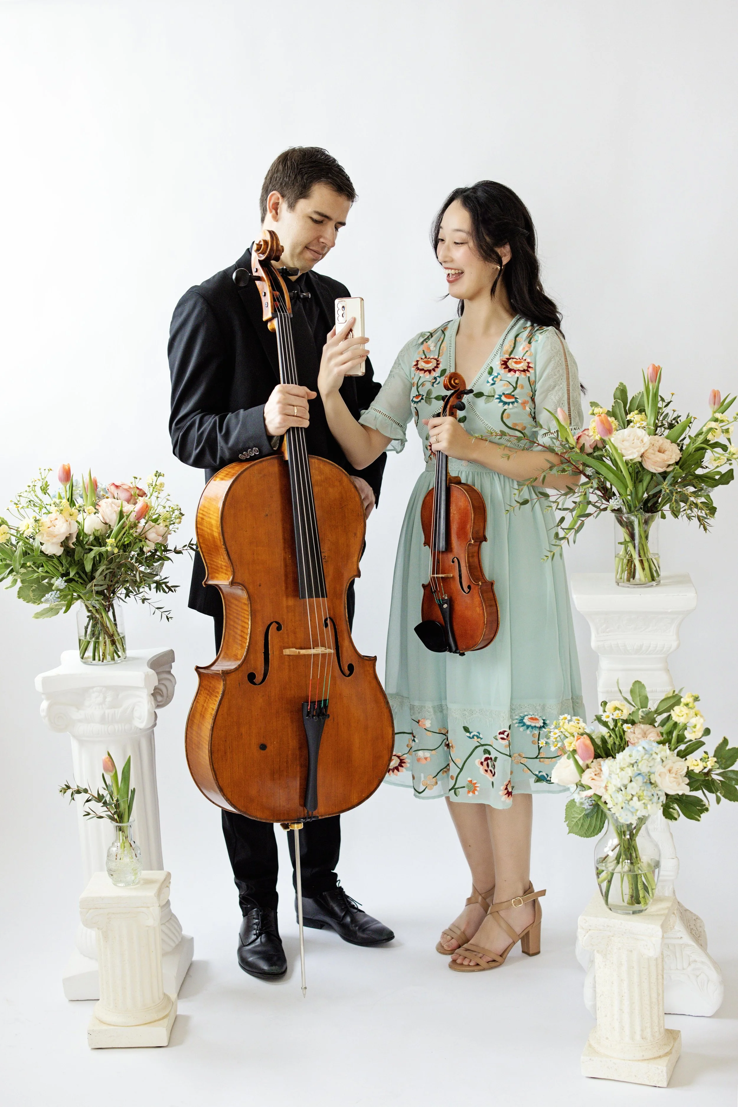 A man and a woman standing together holding musical instruments, surrounded by flower arrangements, smiling and taking a selfie.