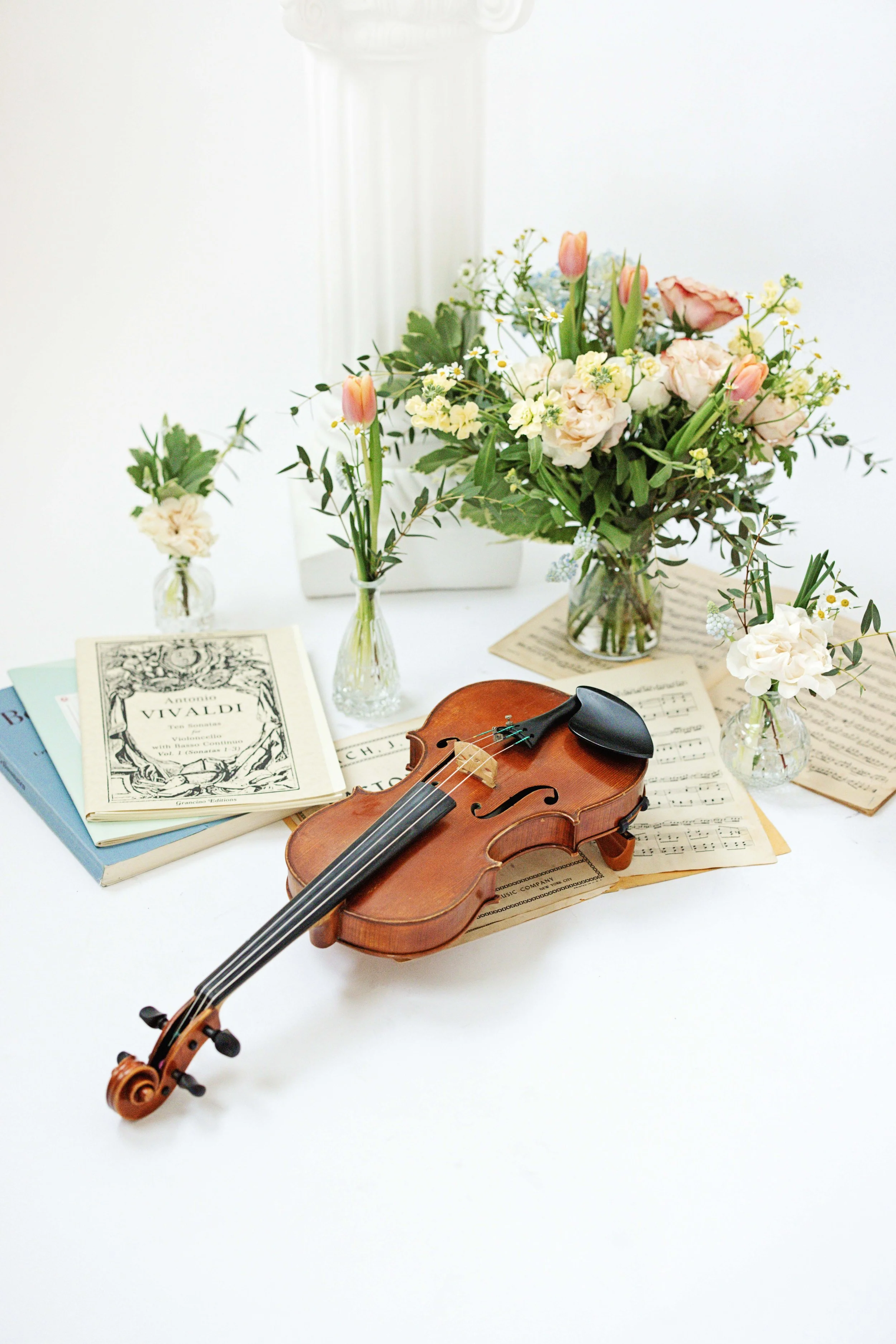 A wooden violin resting on sheet music on a white table, surrounded by open music books and three small vases of pink and white flowers.