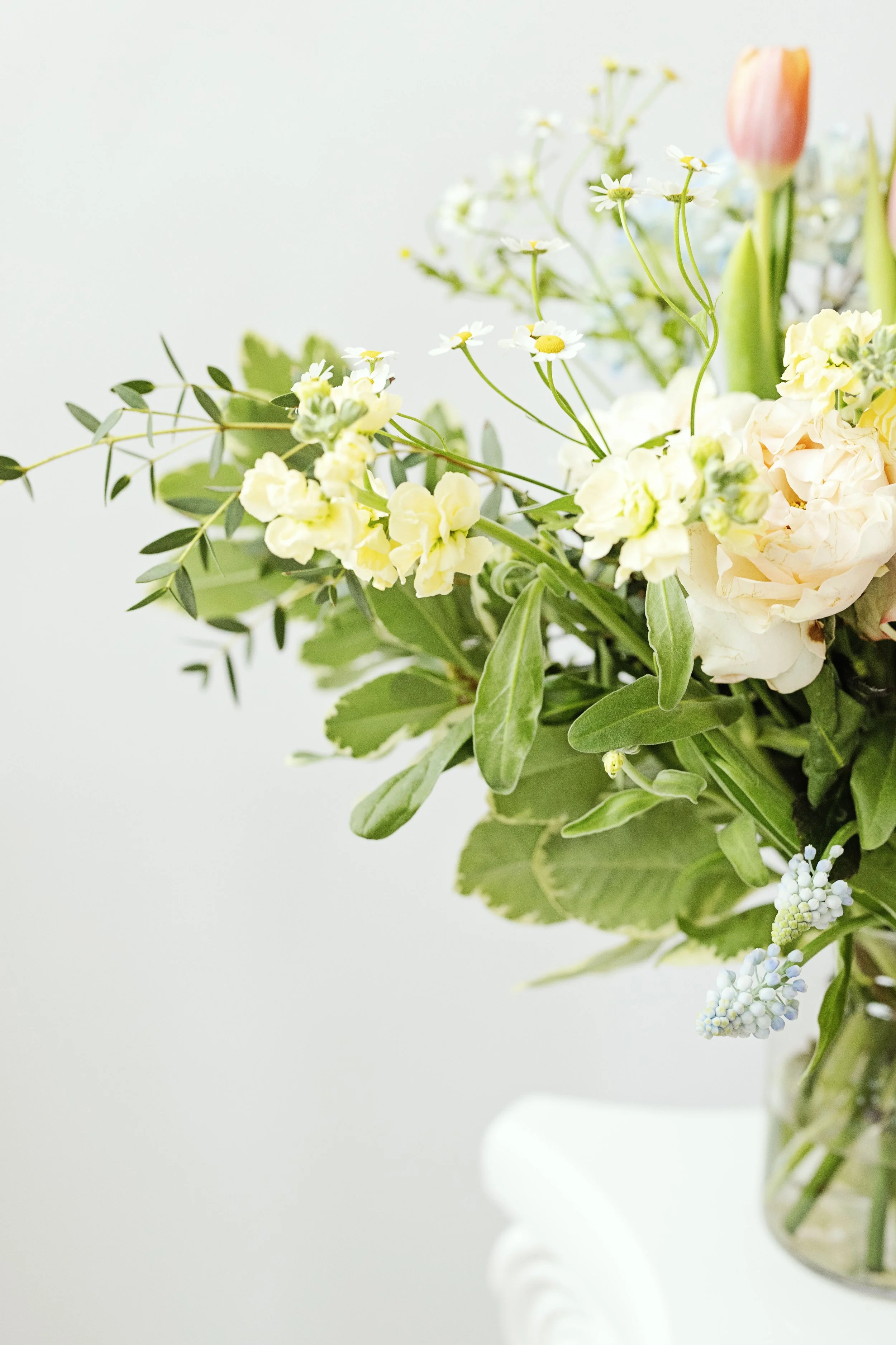 Close-up of a bouquet of assorted flowers in a glass vase, including white, yellow, pink, and green blooms with green leaves against a light background.