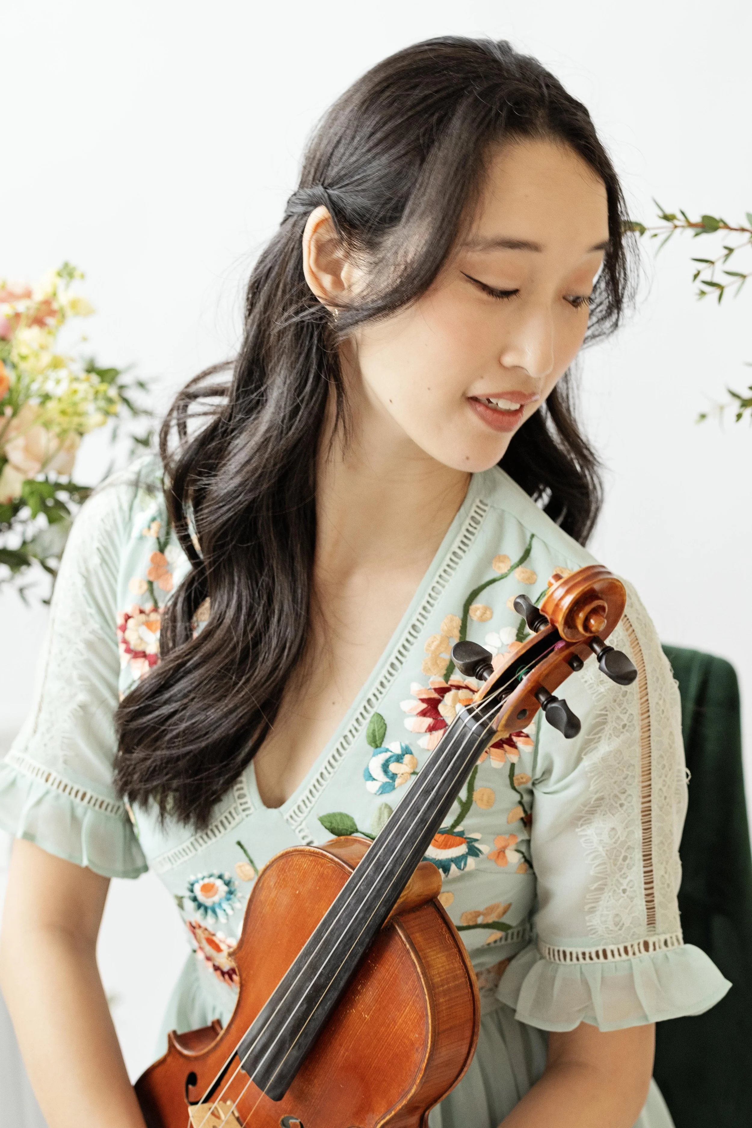 A woman with long dark hair playing a violin, wearing a light-colored embroidered dress, with flowers in the background.