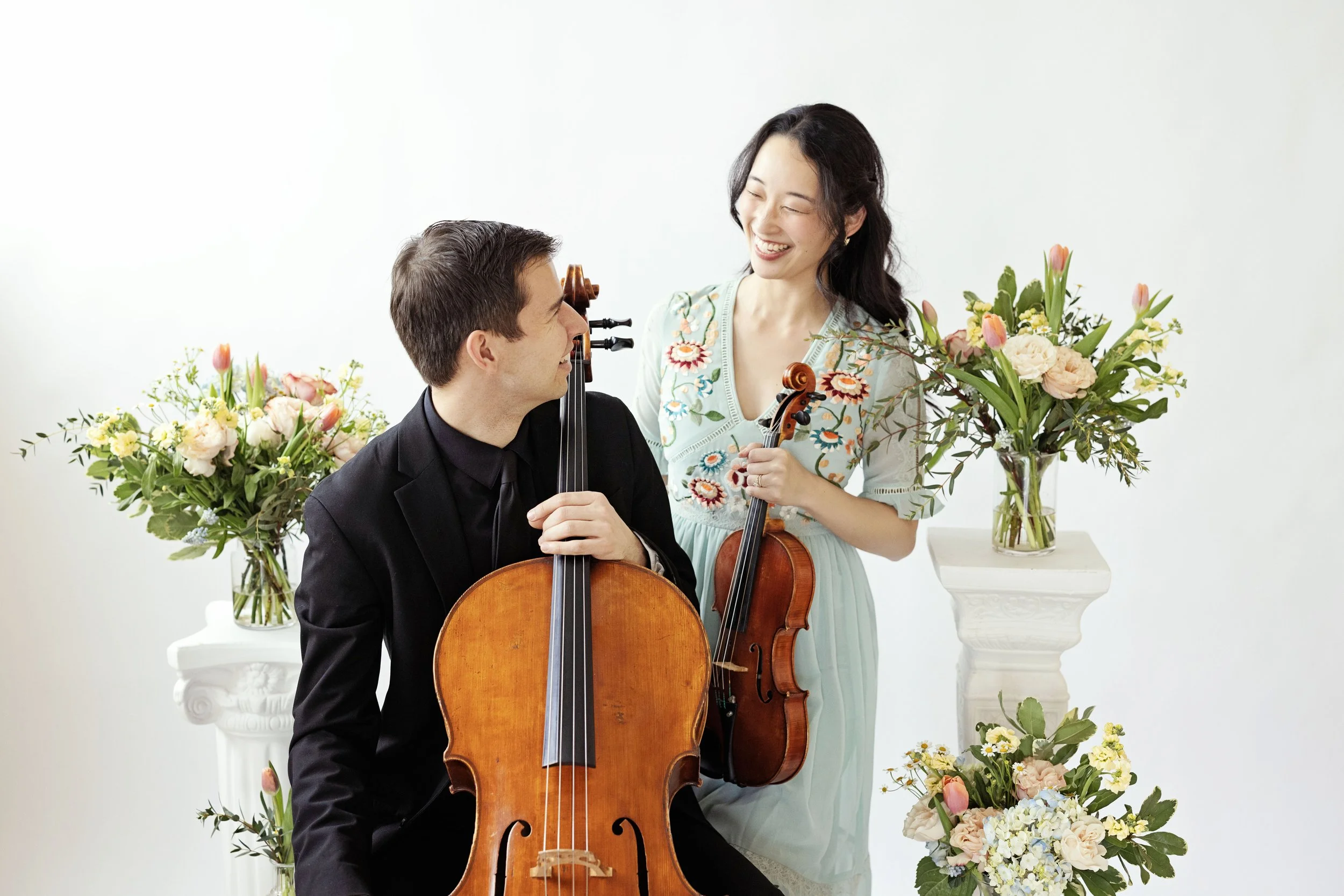 A wedding cellist and violinist smiling at each other in a brightly lit room