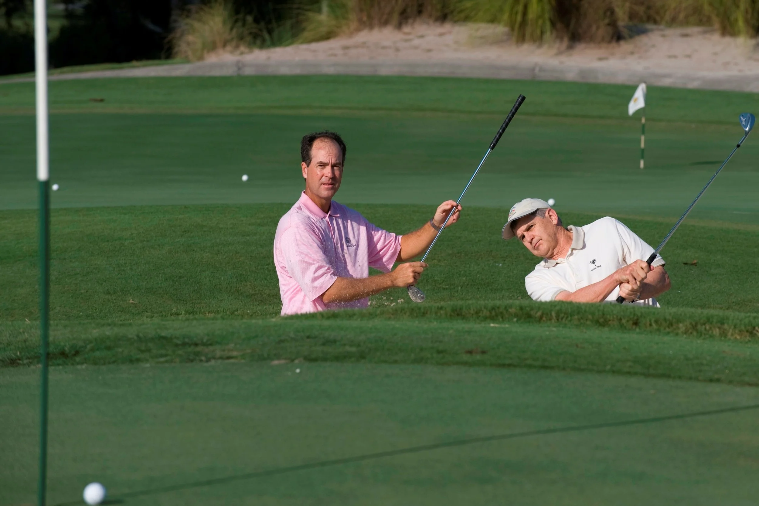 Two men practicing golf on a green golf course, one is standing and the other is in a bunker's sand. Both are holding golf clubs, and golf balls are scattered on the green.