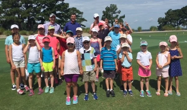 Group of children and adults on a golf course posing for a photo, some children wearing colorful clothing and hats.