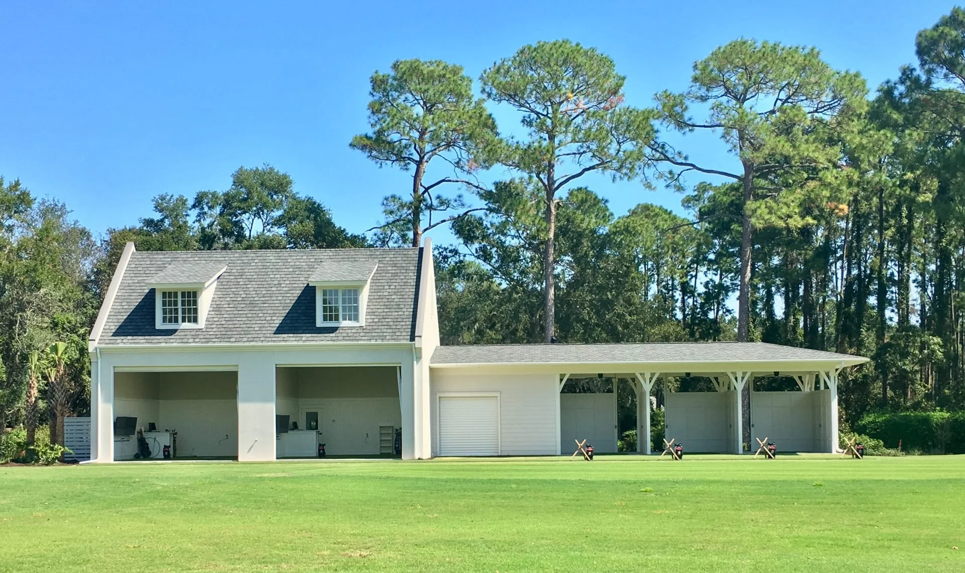 A large, white house with a steep gray roof featuring two dormer windows, surrounded by tall pine trees, with a spacious grassy lawn and a covered outdoor area with golf course markers or signs in the foreground.