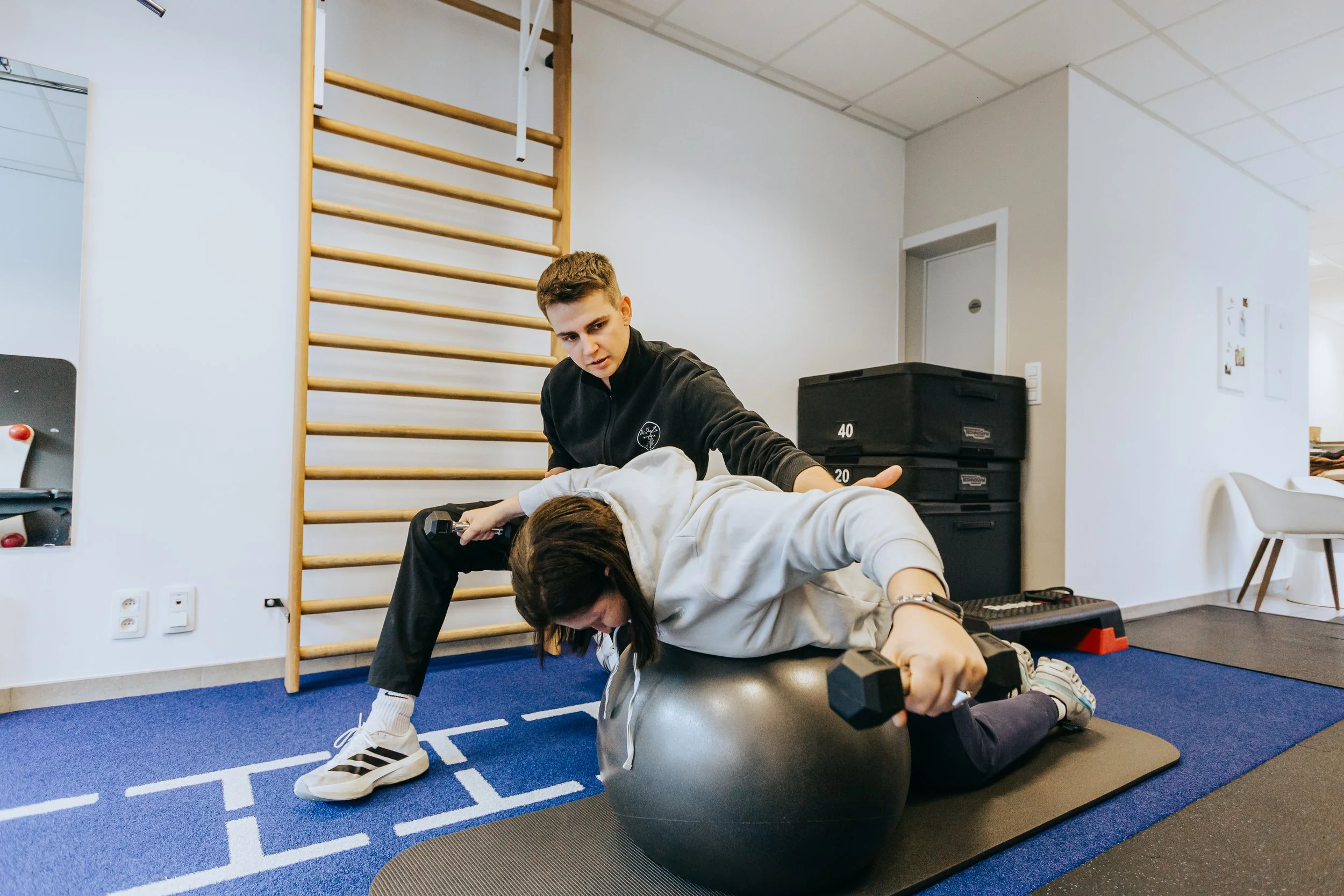Een jonge man helpt een jonge vrouw met fitnessoefeningen, waarbij ze op een medicijnbal liggen en een stretch doen in een sportschool of fysiotherapiecentrum.